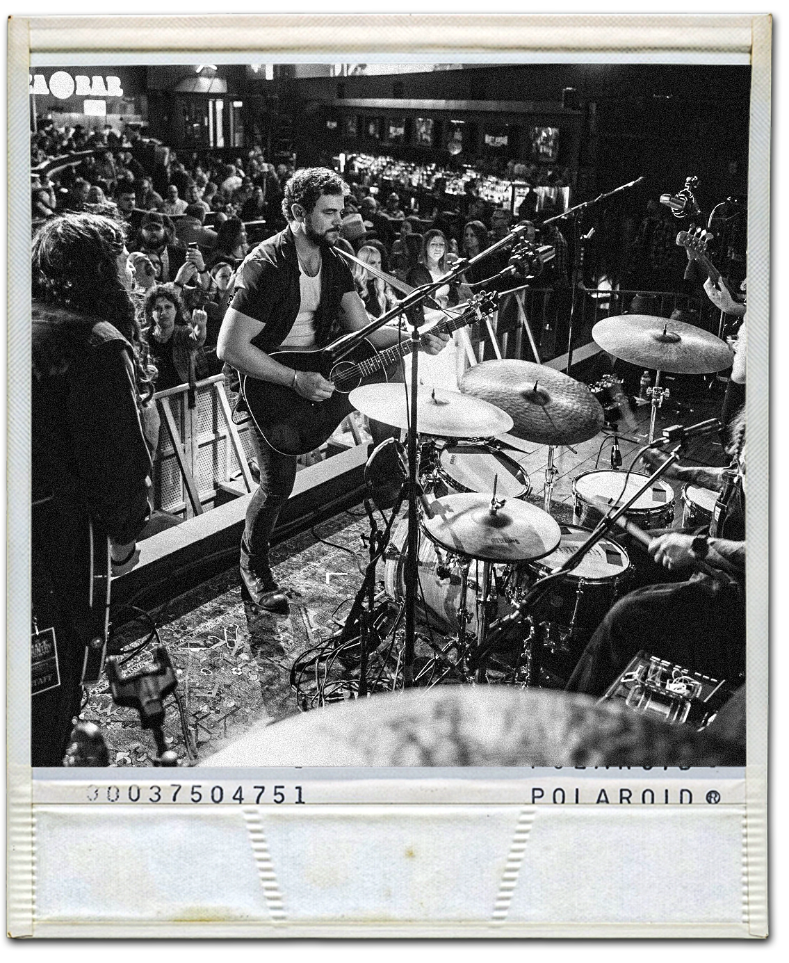 Black and white photo of a live music performance with a guitarist and drummer on stage, audience in the background in a bar or concert venue.