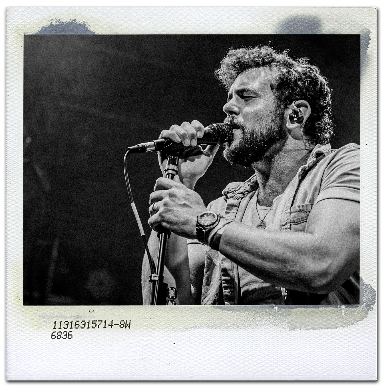 Black and white photo of a man singing into a microphone on stage, wearing a sleeveless shirt, a watch, and a necklace.