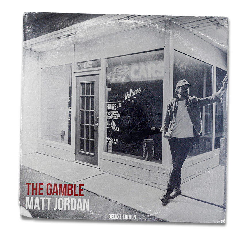 Black and white photo of a man standing outside a storefront with large glass windows and a door, holding a cigarette and leaning against the glass. The store has signage for a car lot and dollar store. Text on the image reads "The Gamble" by Matt Jordan, Deluxe Edition.