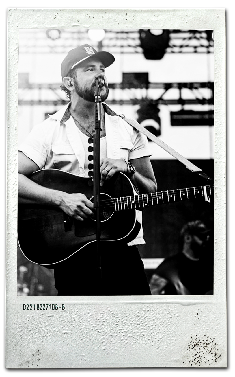 Black and white photo of a male musician with a beard singing into a microphone while playing an acoustic guitar on stage. He is wearing a cap, a short-sleeve shirt, and a necklace. A man with glasses is visible in the background.
