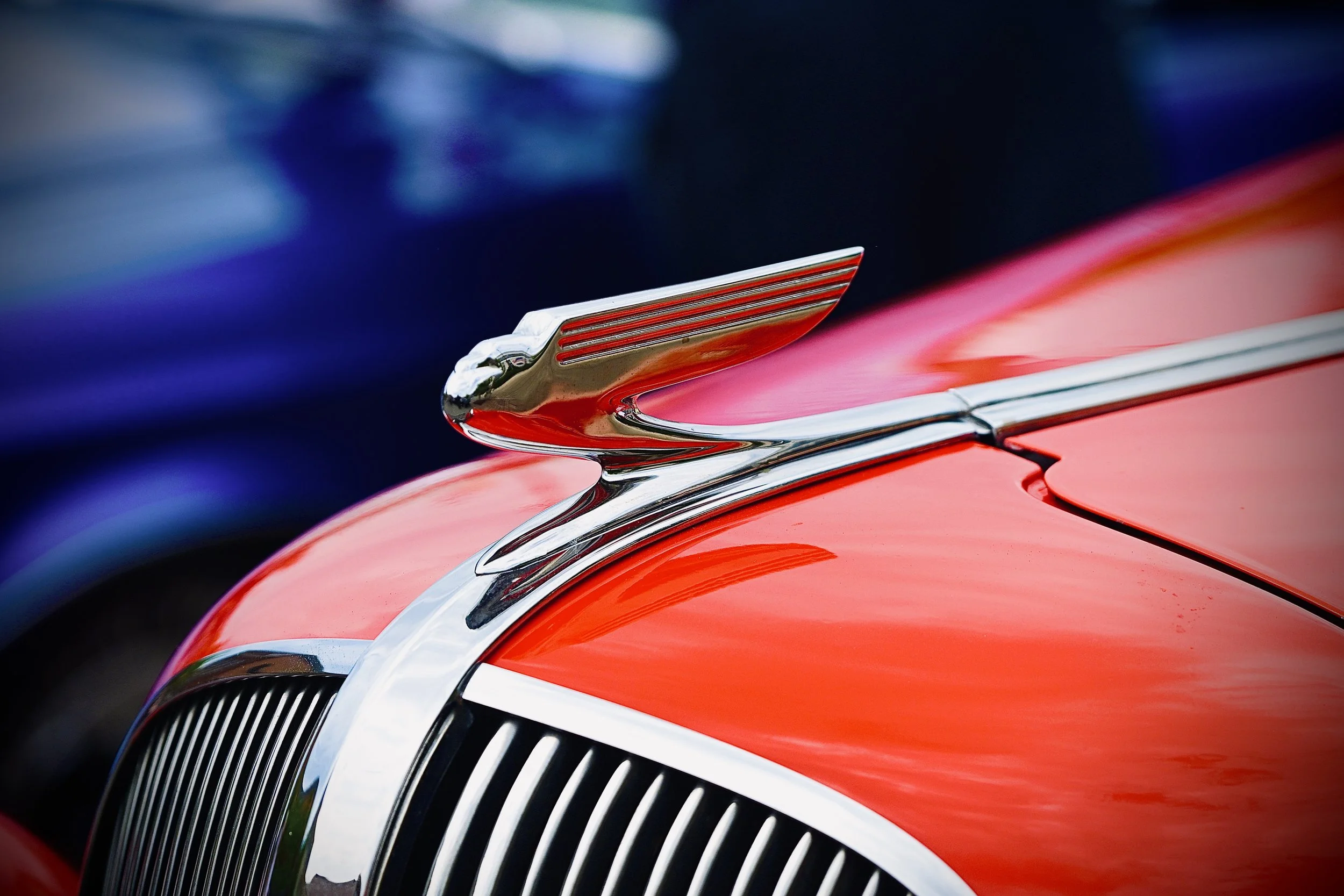 Close-up of a vintage car's hood ornament with red and chrome detailing, red paint, and a black background.