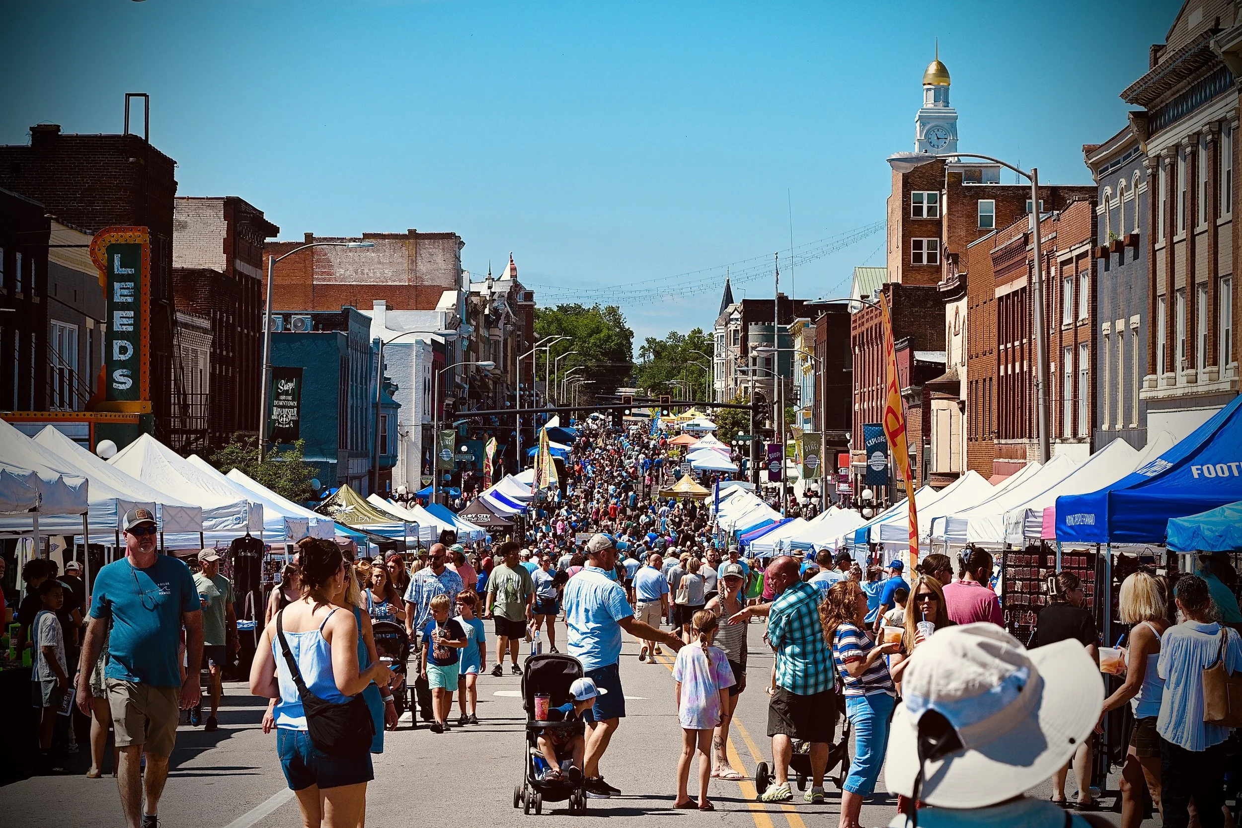 Crowd of people walking and shopping at the Beer Cheese Festival with food and vendor tents, city buildings, and a clock tower in the background on a sunny day.