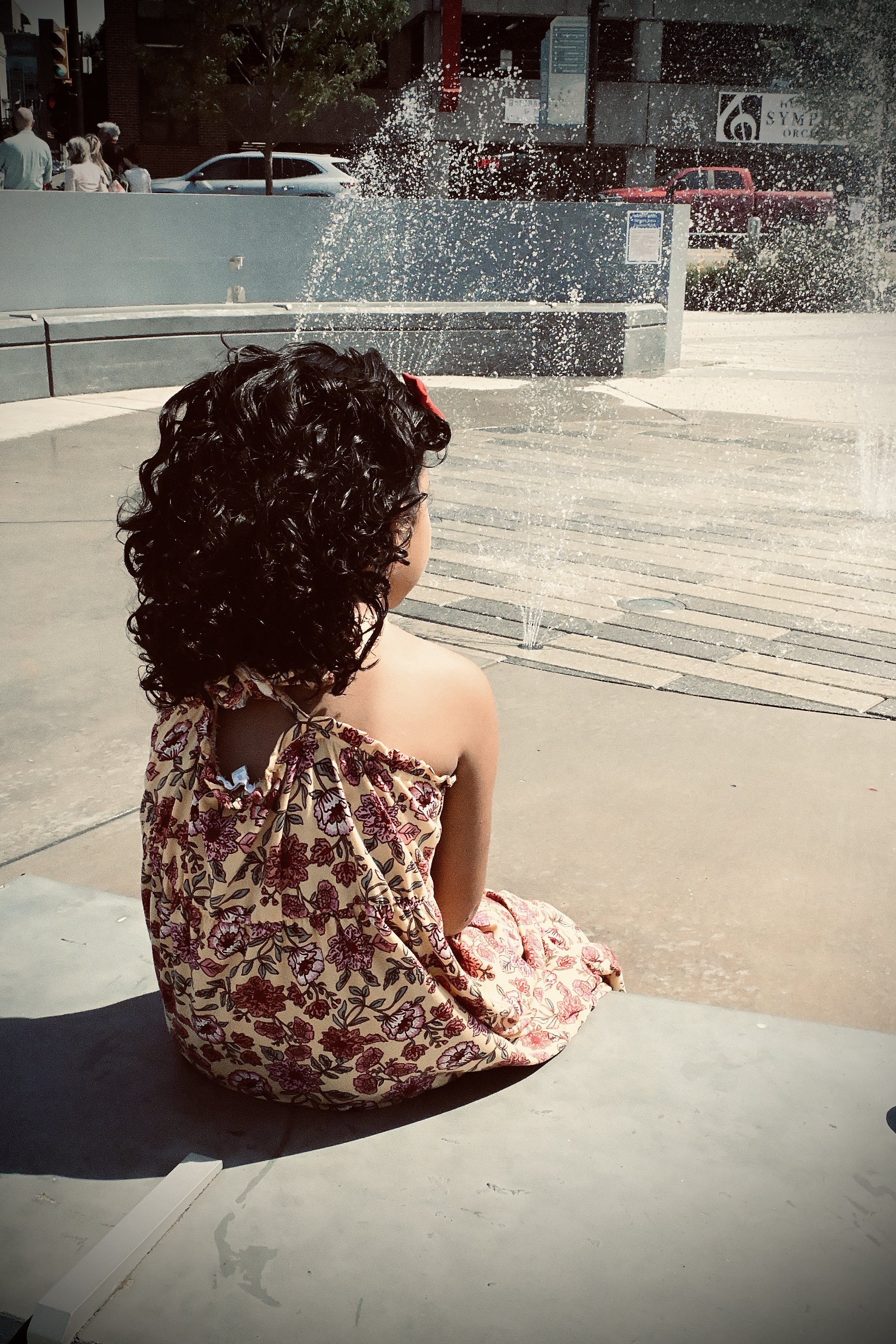A young girl with curly dark hair, wearing a floral dress with red and pink flowers, sitting on a concrete surface near a water fountain in an urban area, with people walking and cars visible in the background.