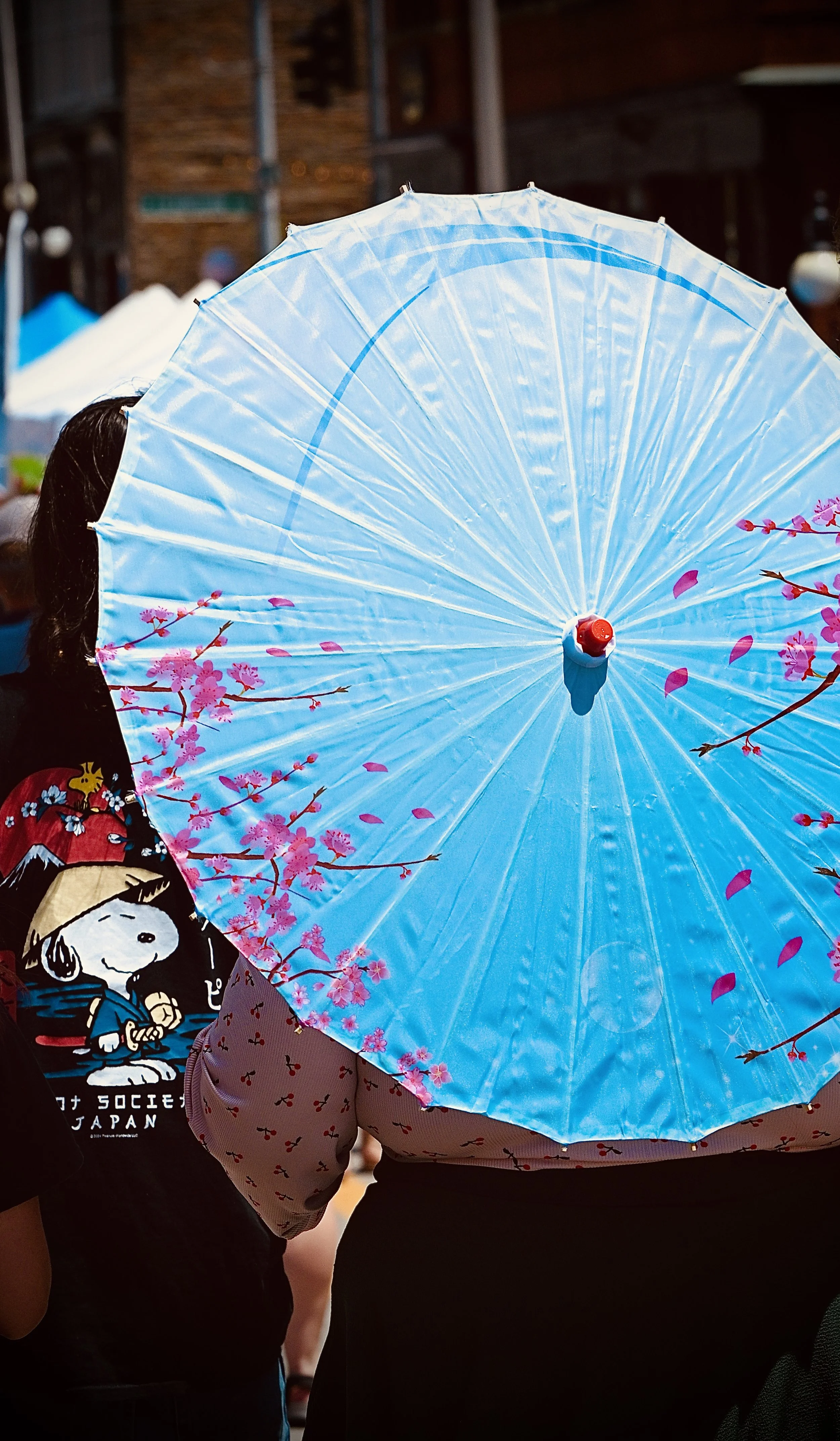 People in a crowded outdoor setting, one person holding a blue parasol with pink cherry blossom designs, and one wearing a black shirt with Snoopy from Peanuts.