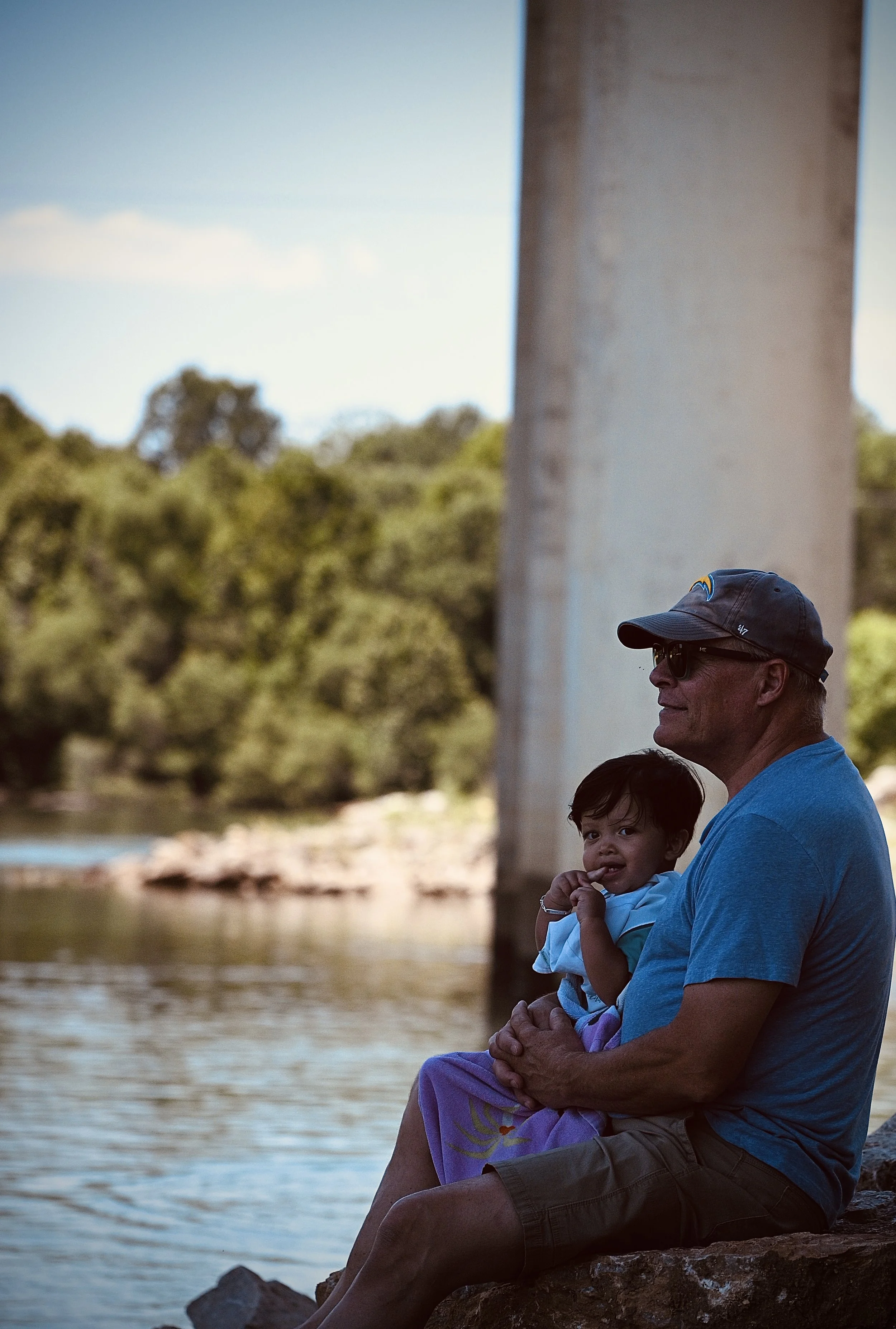 An older man with sunglasses and a cap sitting on rocks near a river, holding a young child in his lap. They are outdoors with trees and water in the background.