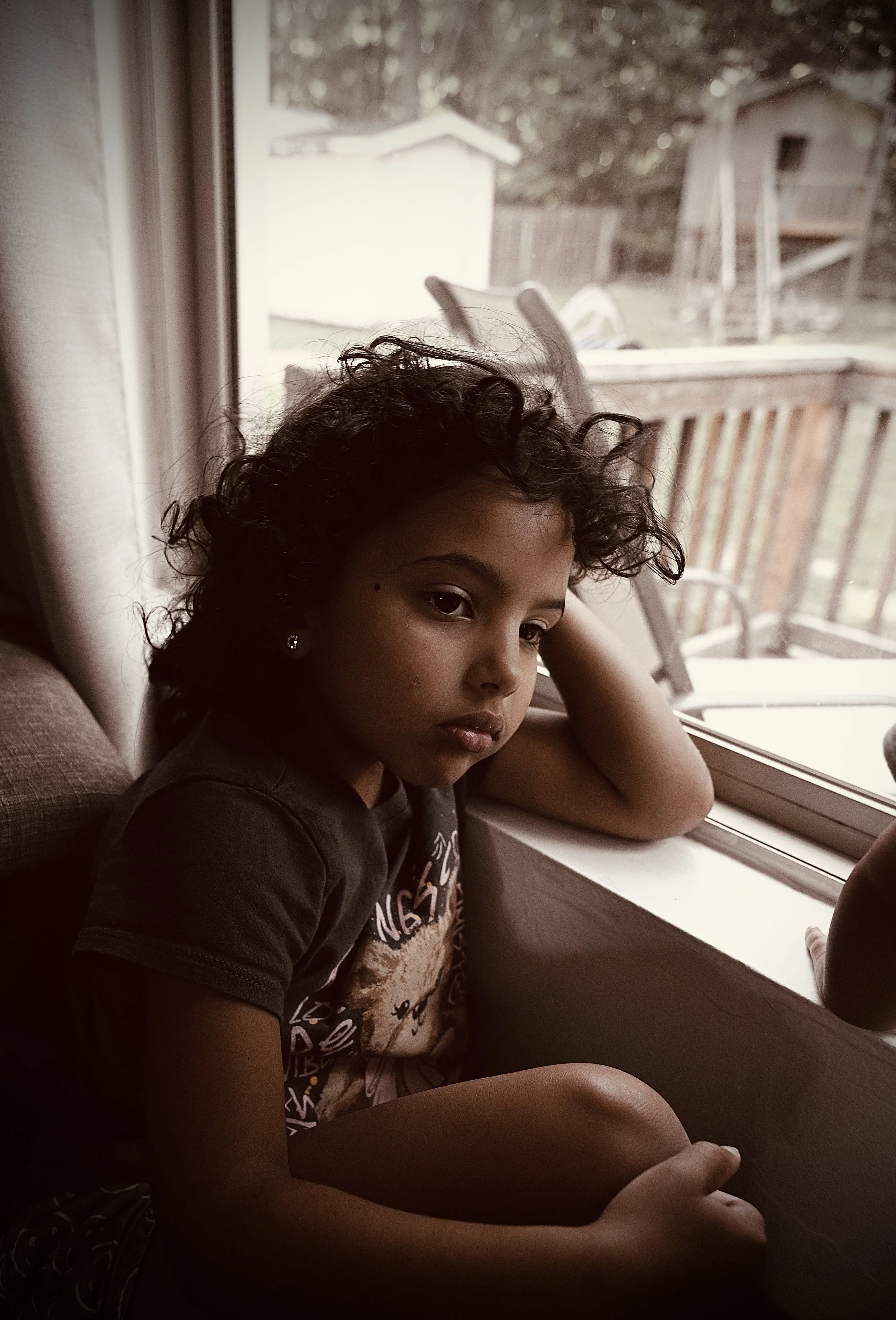 A girl with curly hair and earrings sitting by a window, looking outside with her arm resting on the window sill. Nearby, there is a wooden deck and shed outside.
