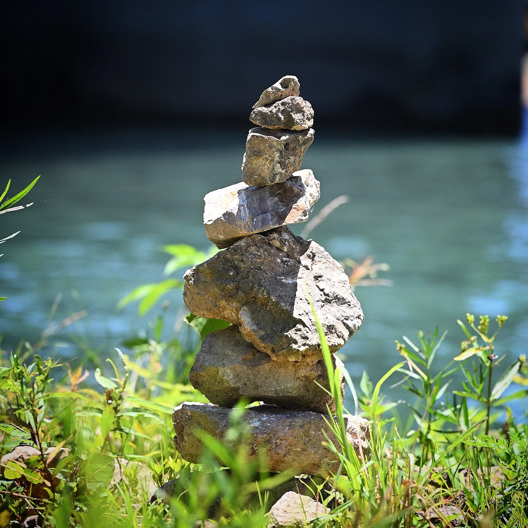 A stack of seven rocks balanced on top of a larger rock near a body of water with greenery in the foreground.