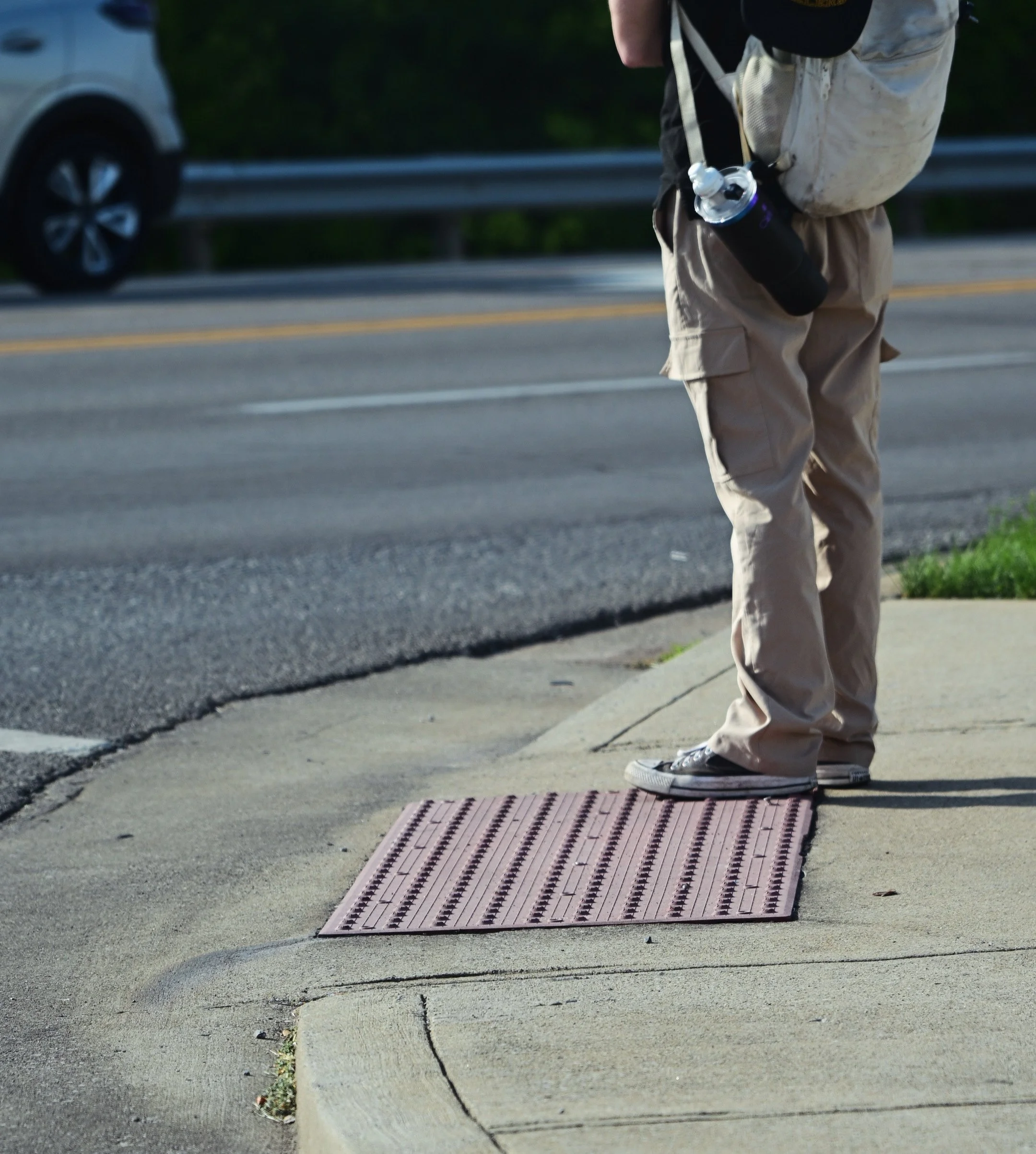 A person standing on a sidewalk near a street, wearing beige cargo pants and grey shoes, with a backpack and carrying a water bottle. A grey car is parked on the street.