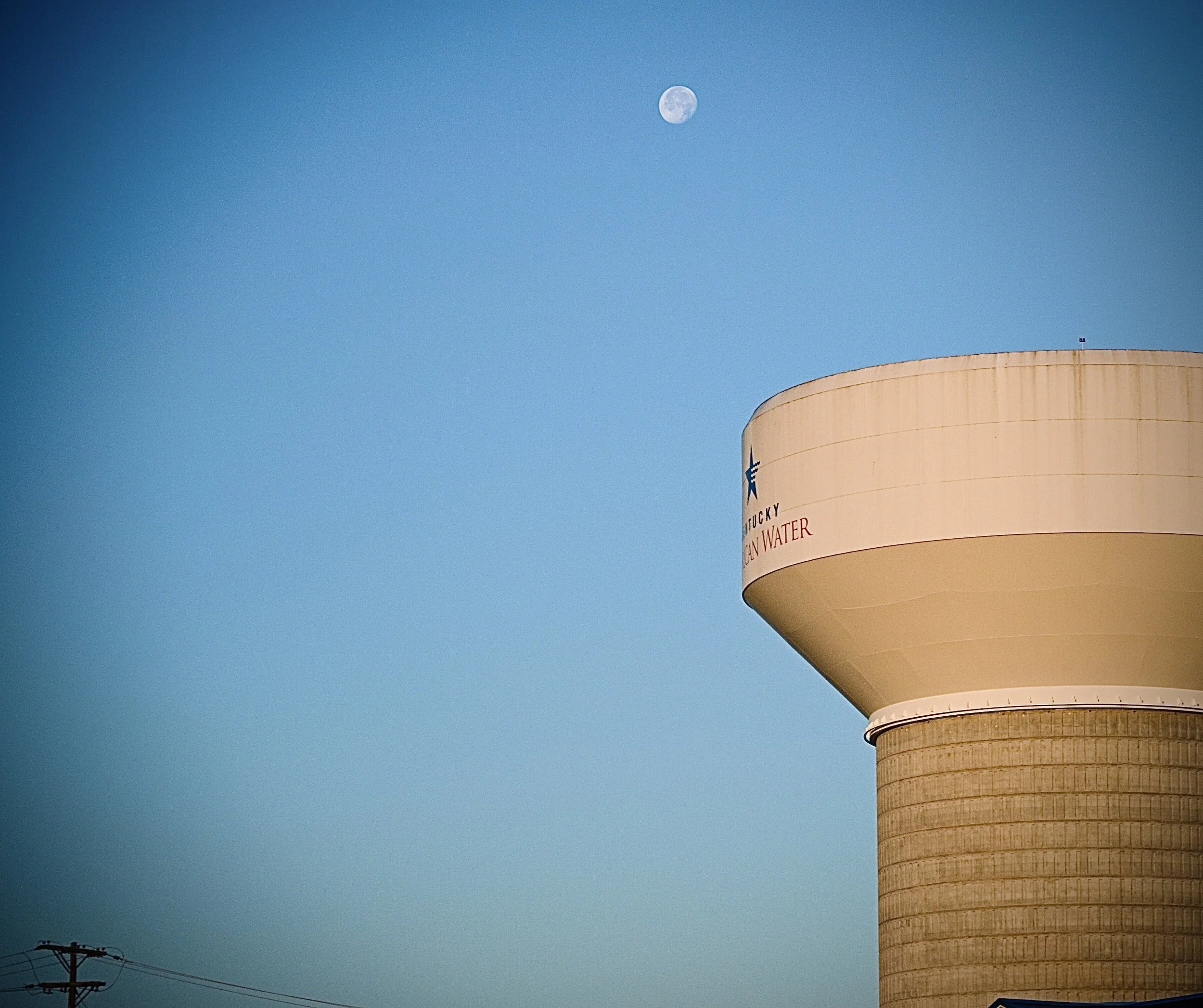 Near the top of a tall water tower with a beige exterior and a rounded shape, the moon is visible in a clear blue sky. Power lines and poles are seen at the bottom left.