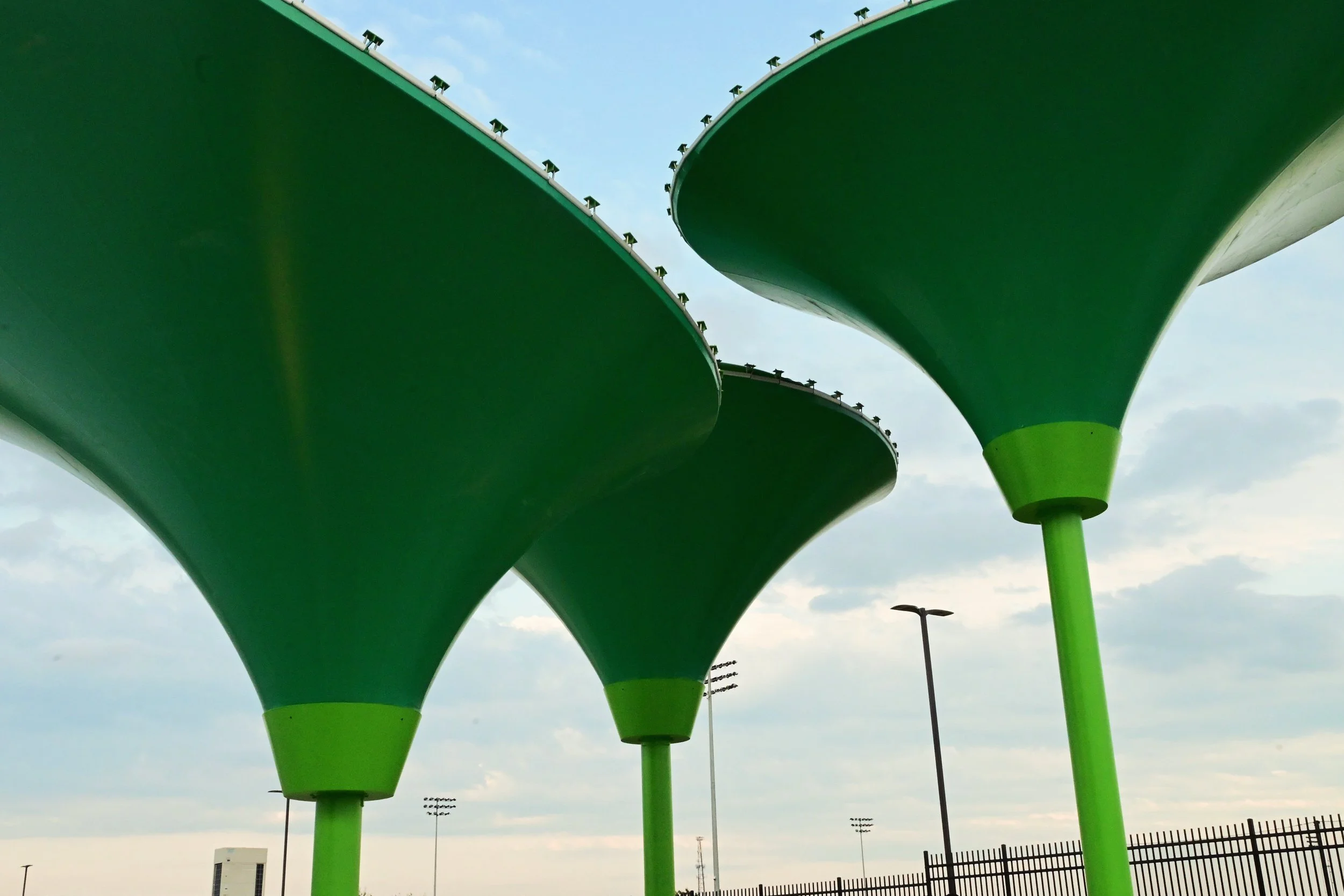 Large green abstract structures elevated on green poles, under a partly cloudy sky. At Lexington Sporting Club Field