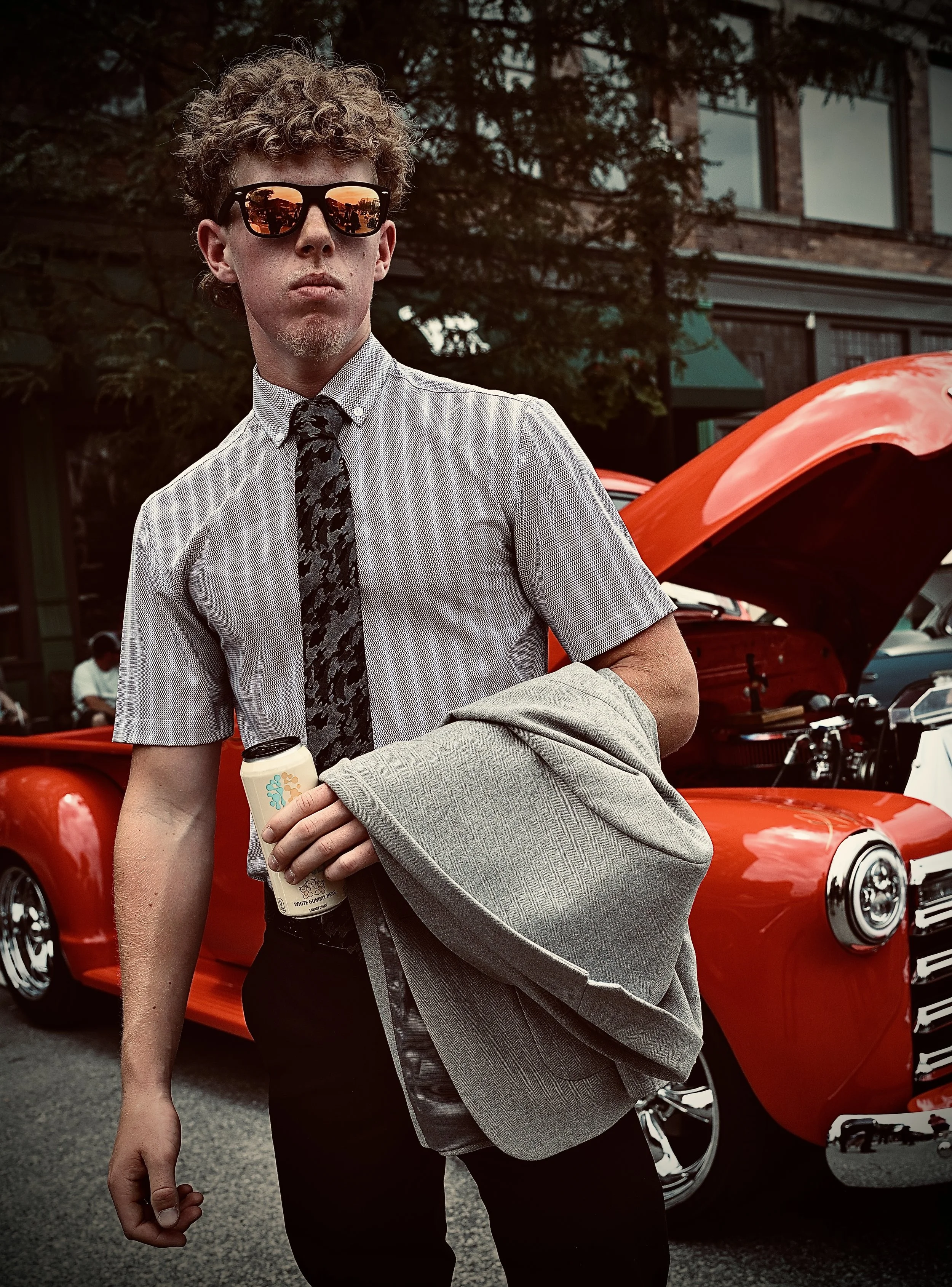 A young man wearing sunglasses standing in front of a red vintage truck, holding a drink and a gray jacket, in an outdoor urban setting.