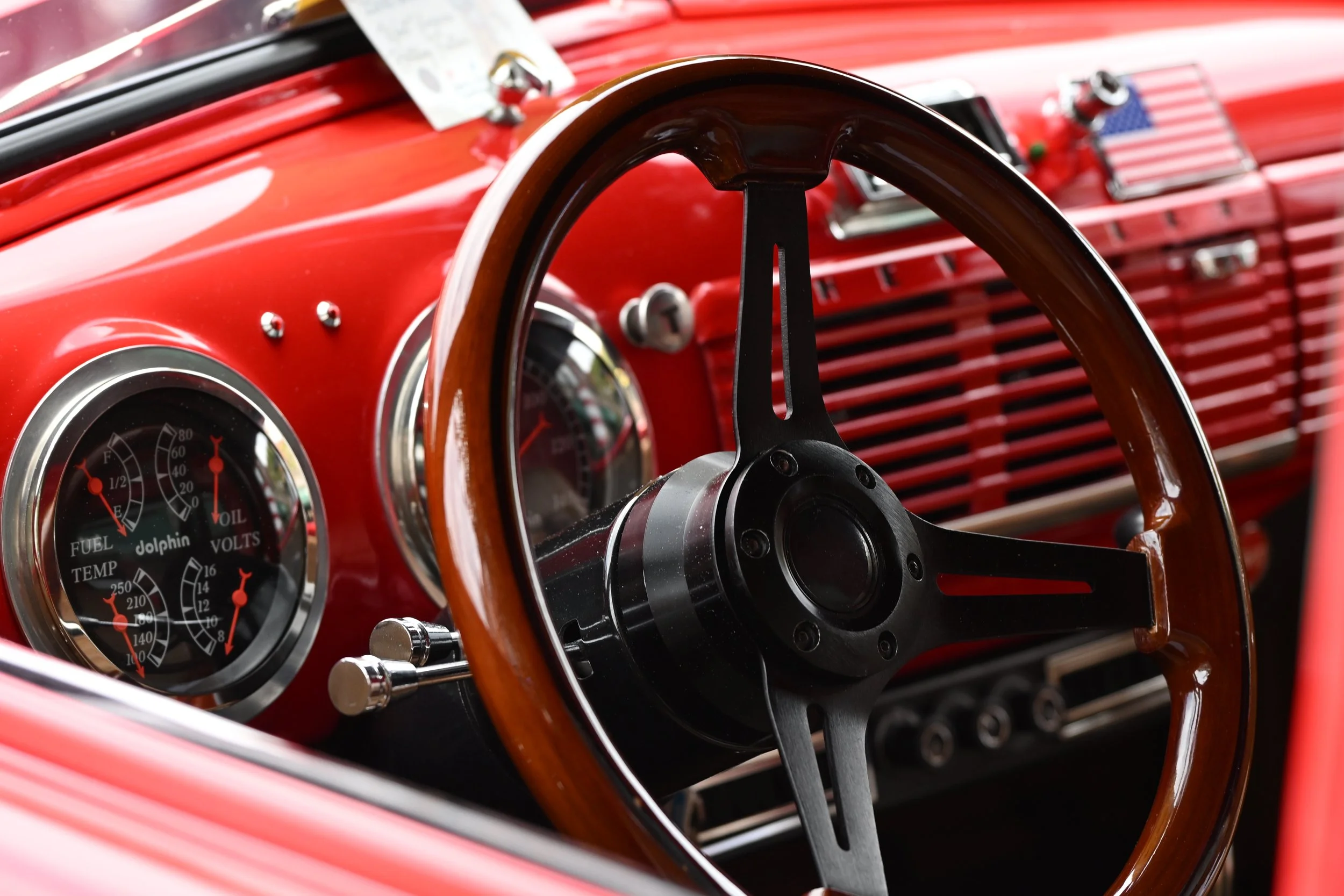 Close-up view of the interior of a vintage red car, showing the steering wheel made of wood and metal, dashboard gauges including fuel, temperature, volt, and oil pressure meters, and a glimpse of the front grille with an American flag decal.