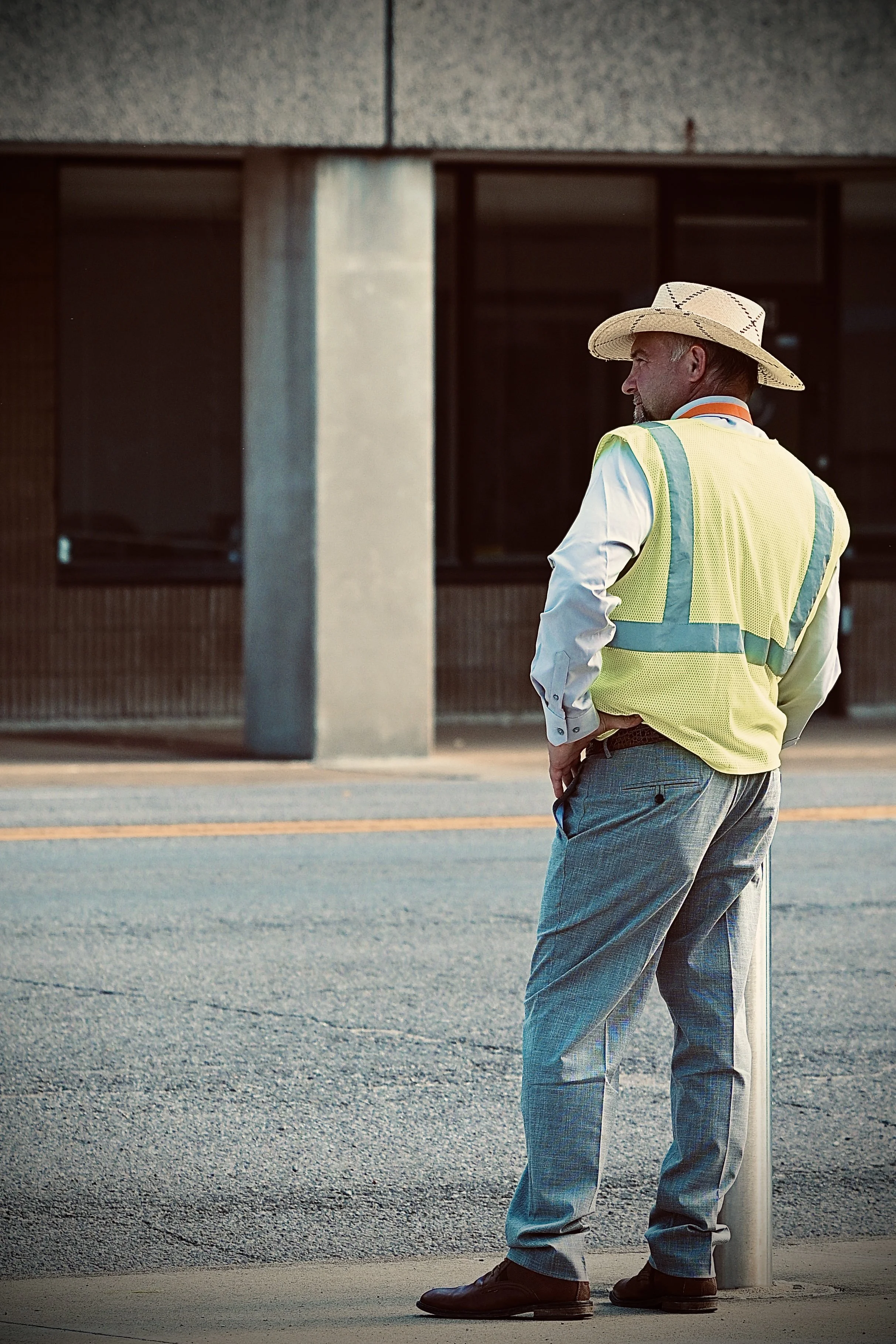A man wearing a wide-brimmed straw hat, yellow safety vest, white shirt, gray trousers, and brown shoes standing next to a street pole in an urban area during daylight.