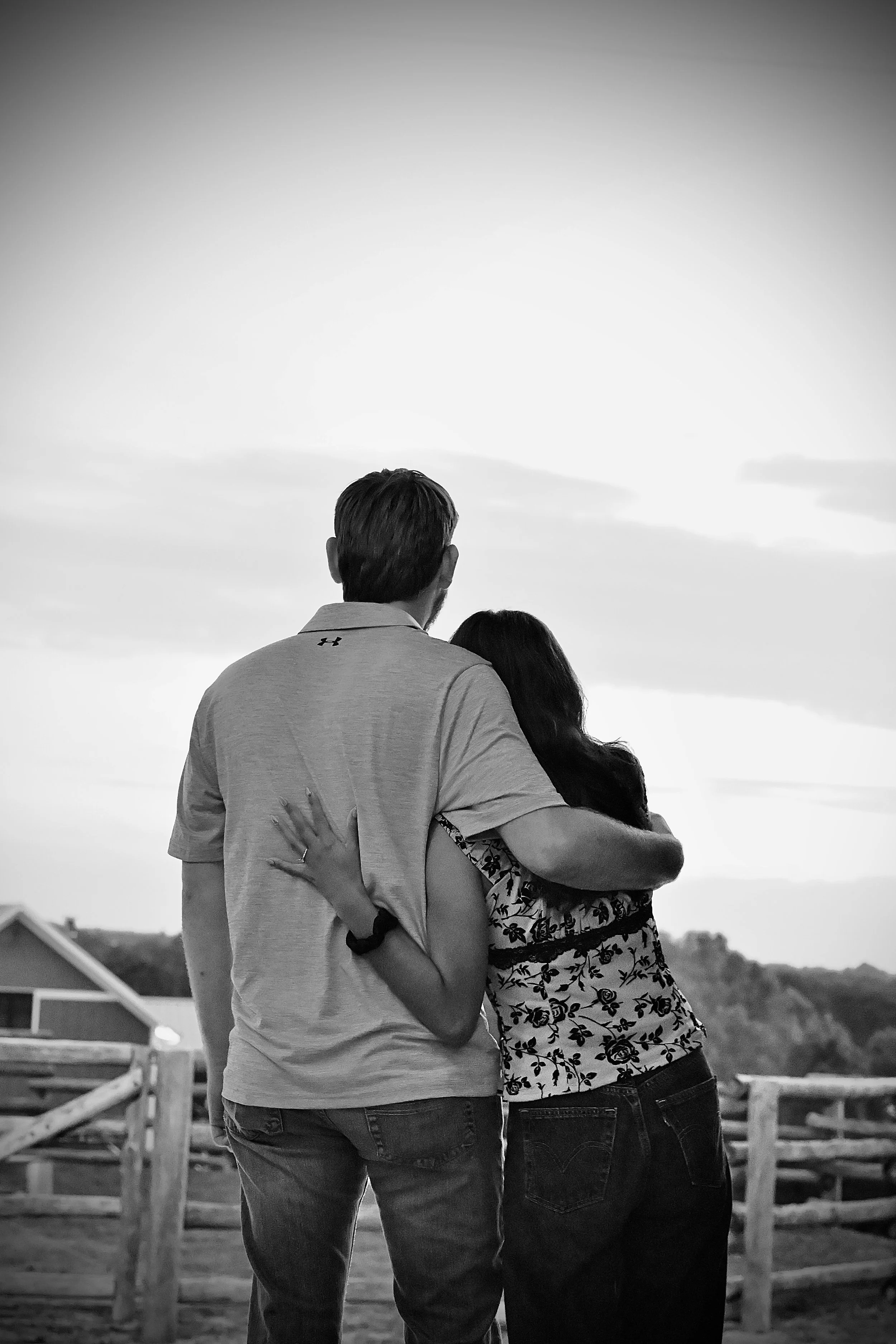 A black-and-white photo of a couple standing outdoors in front of a horse fence, embracing each other, with their back facing the camera as they look at the sky.
