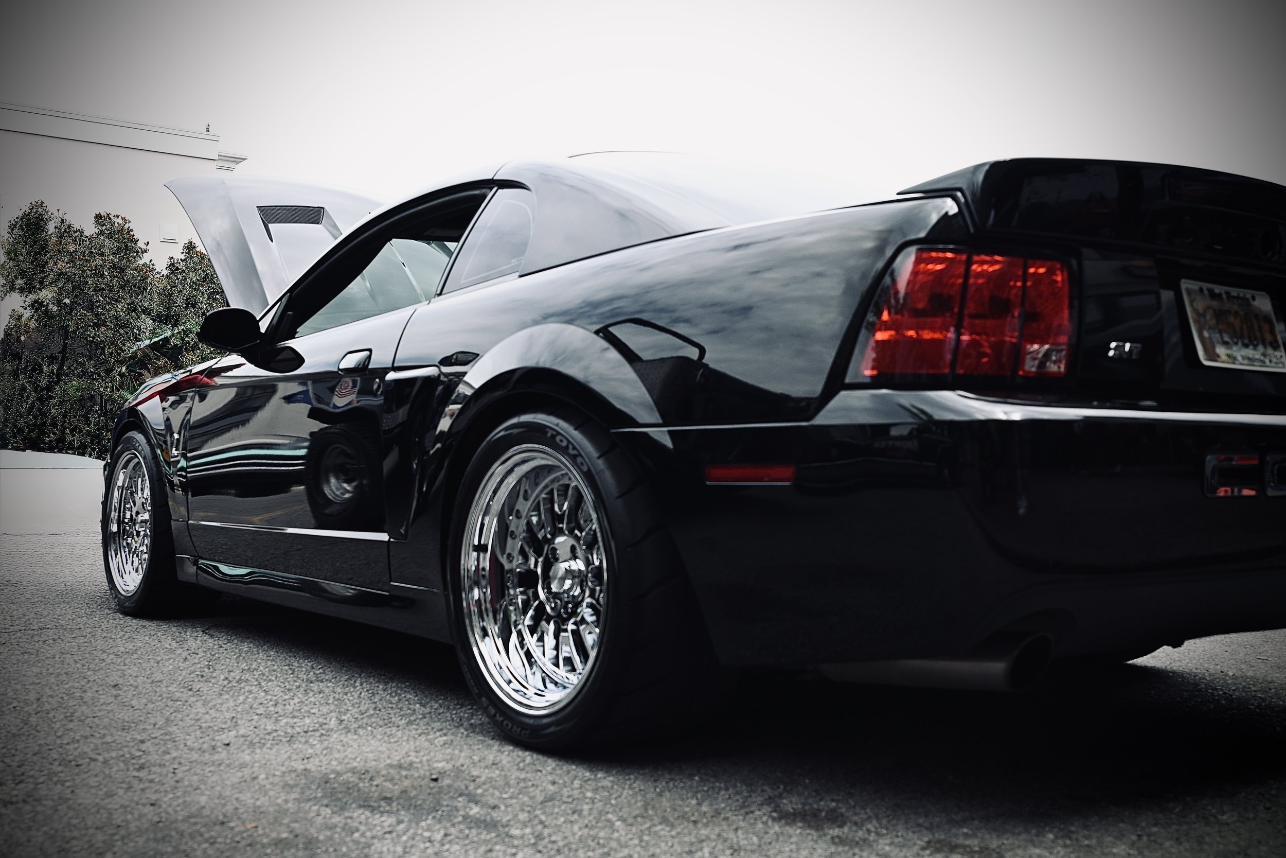 A black sports car with chrome wheels parked on the street, with the hood open and a white building and trees in the background.