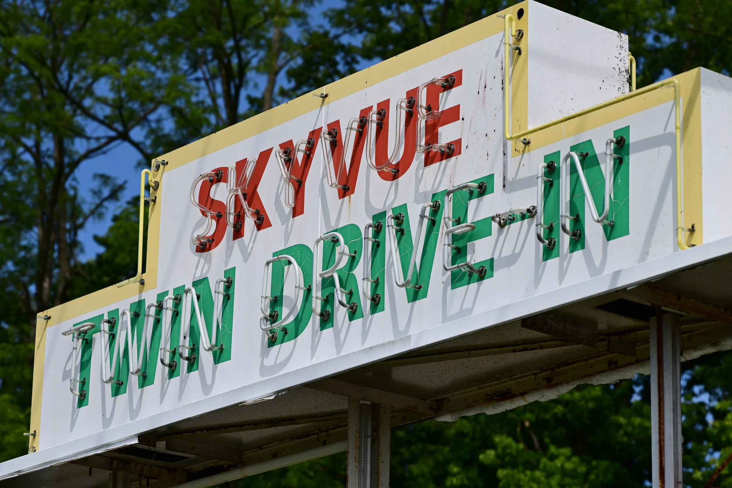 A large sign with neon lights spelling out 'Skyvue Twin Drive-In' , situated outdoors with trees in the background.