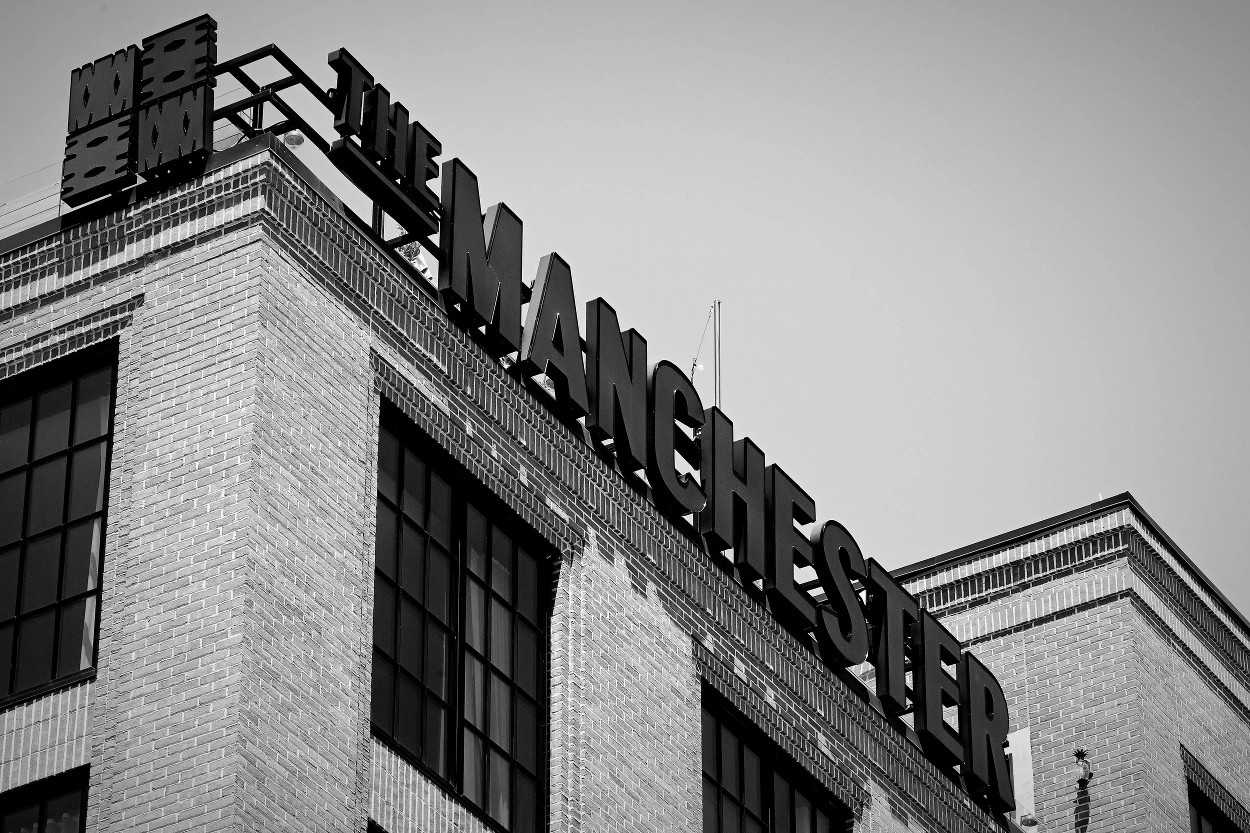 Close-up of a brick building with black-framed windows and a large sign on the roof that reads "THE MANCHESTER" in bold letters, with part of the sign extending beyond the building's edge, in black and white.