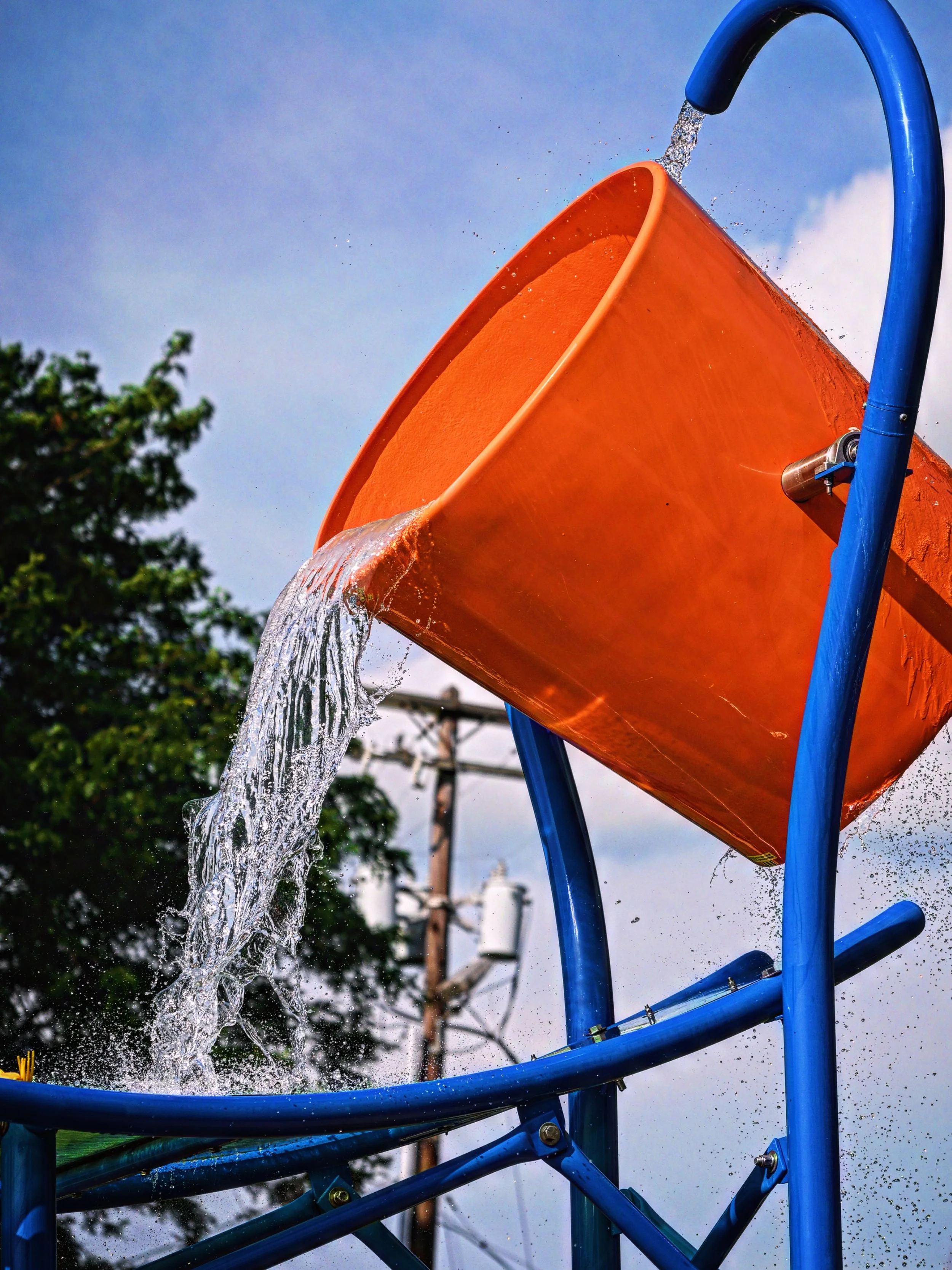 A playground water feature with water flowing out of an orange bucket at the top, water splashing as it falls down, with trees and power lines in the background.