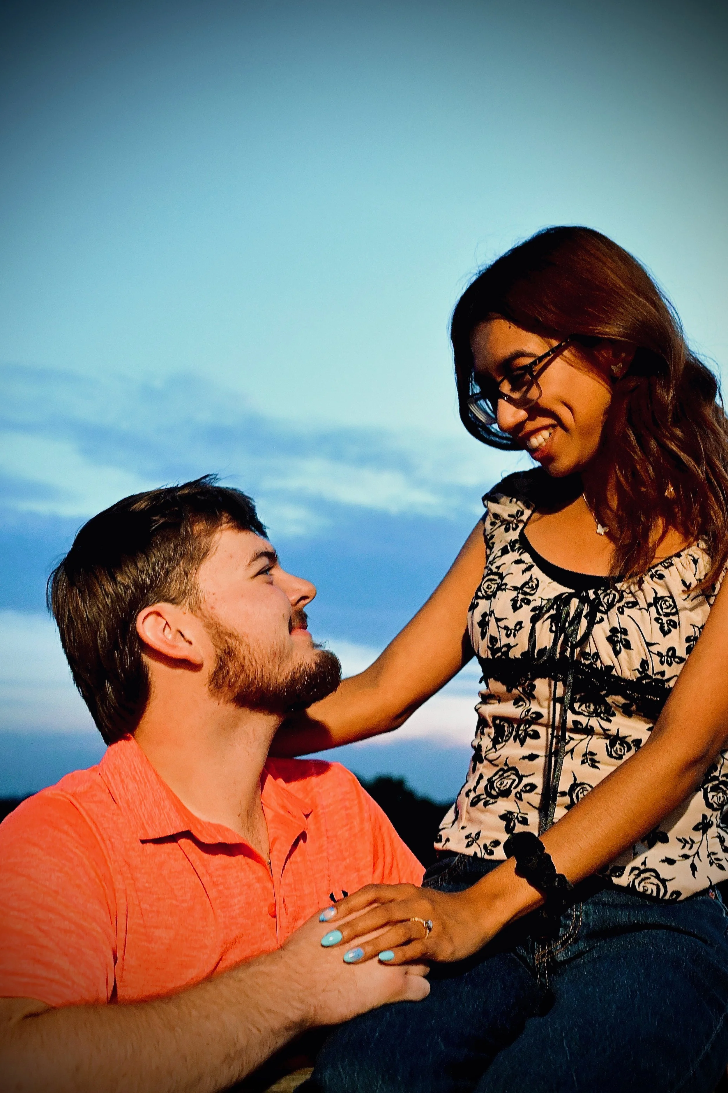 A woman with glasses and long hair smiling at a man with a beard and short hair, who is looking up at her. They are outdoors during sunset with a blue sky and clouds in the background. She said yes!