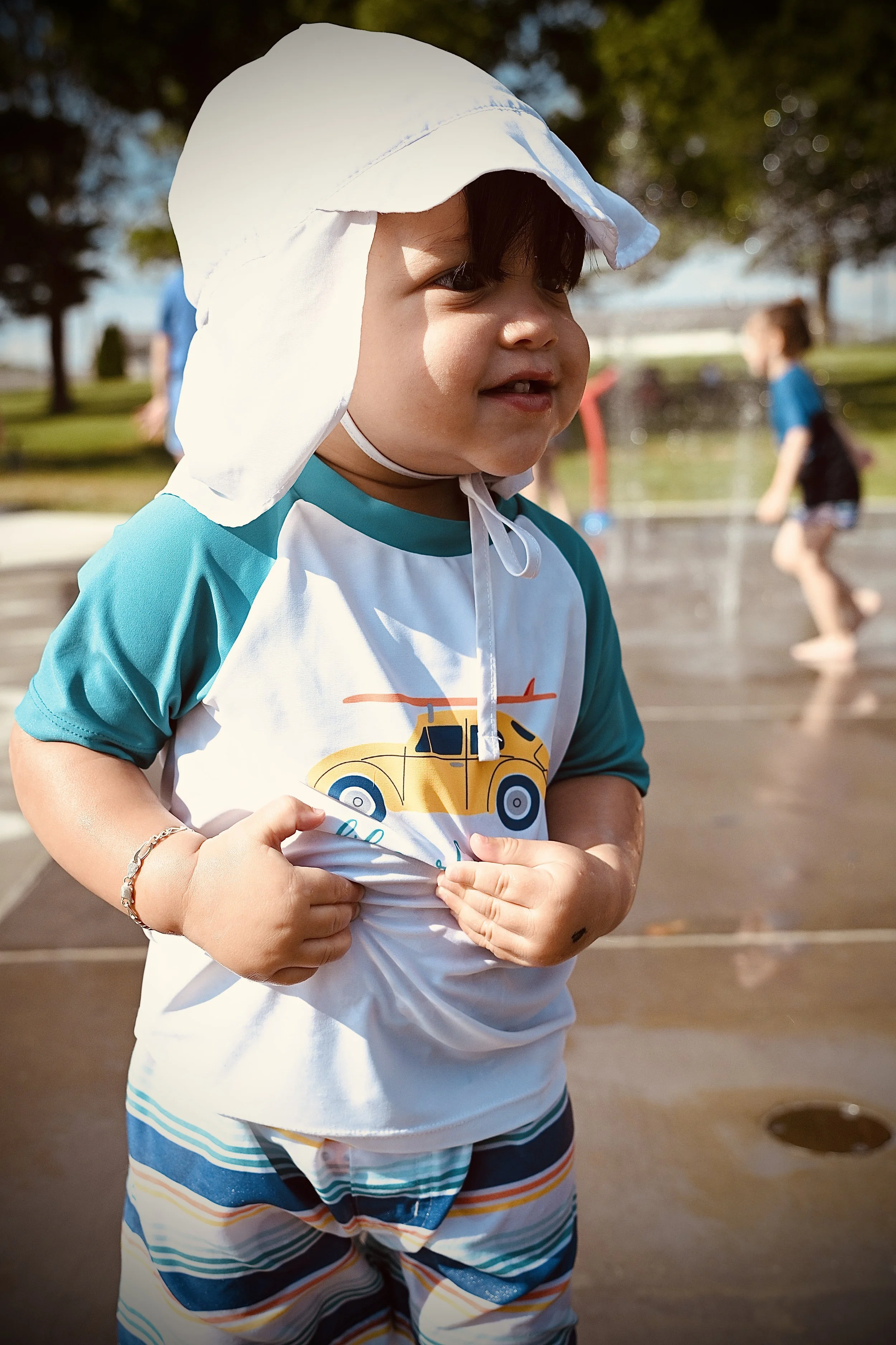 Close-up of a young boy with dark hair, wearing a white sun hat, a T-shirt with a car graphic, and striped shorts, standing outdoors at a splash pad on a sunny day.