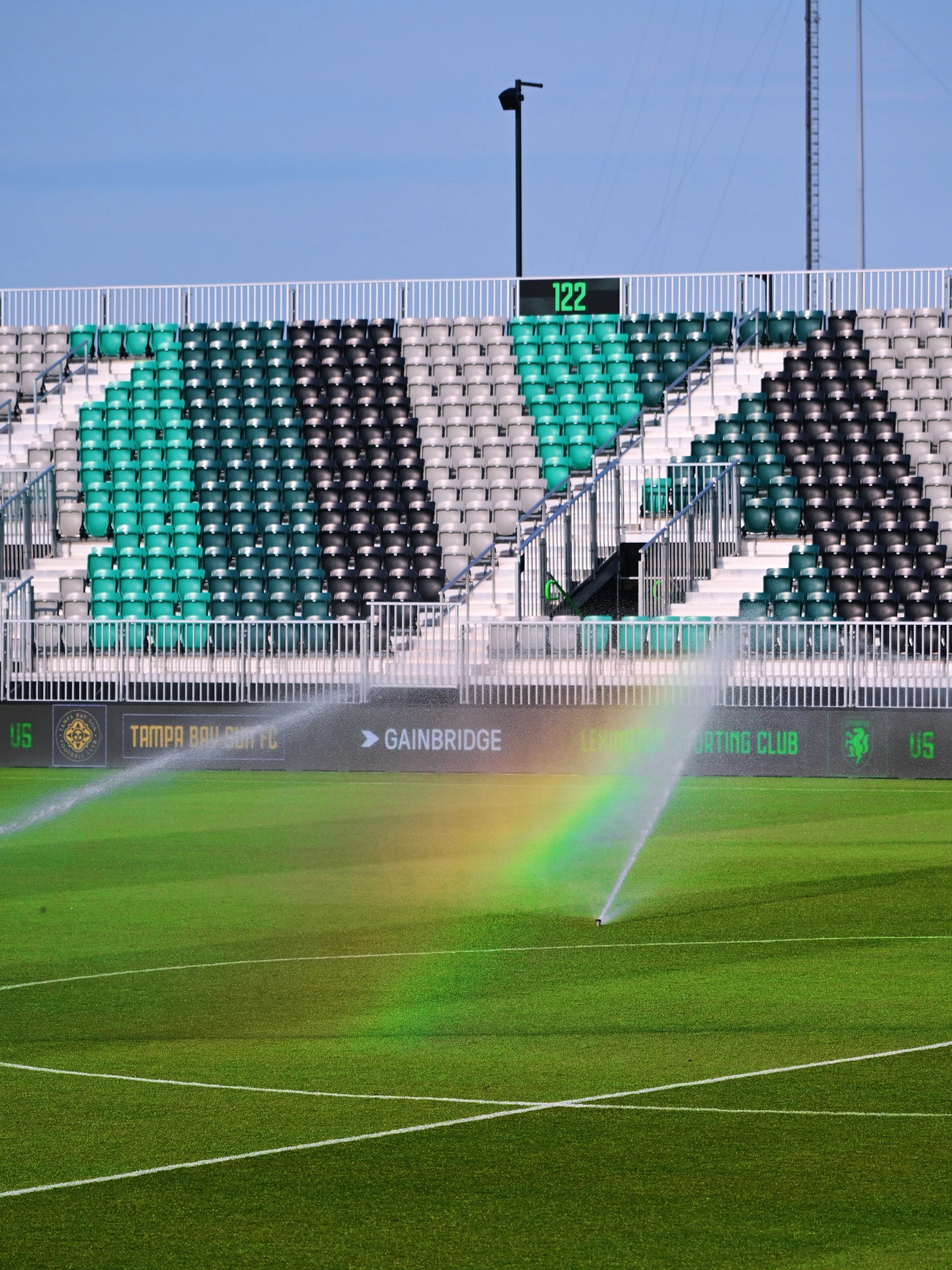 Empty green and black stadium seats in stands above a well-manicured soccer or football field being watered by sprinklers.
