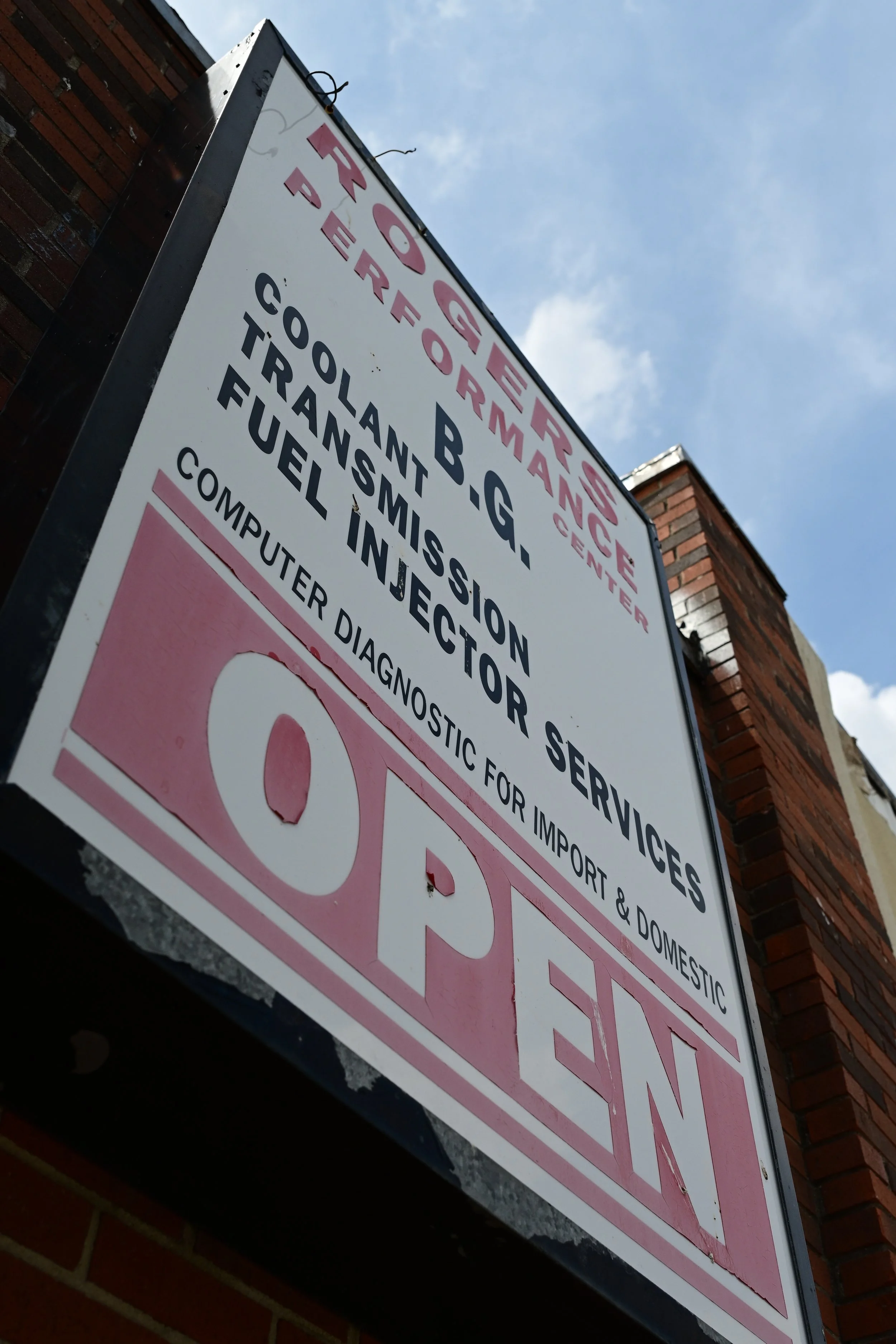 A sign mounted on a brick wall indicates services. The sign lists services like coolant, coolant transfer, and fuel injection, and mentions the availability of computer diagnostic for import and domestic cars. The sky is partly cloudy in the backgrou