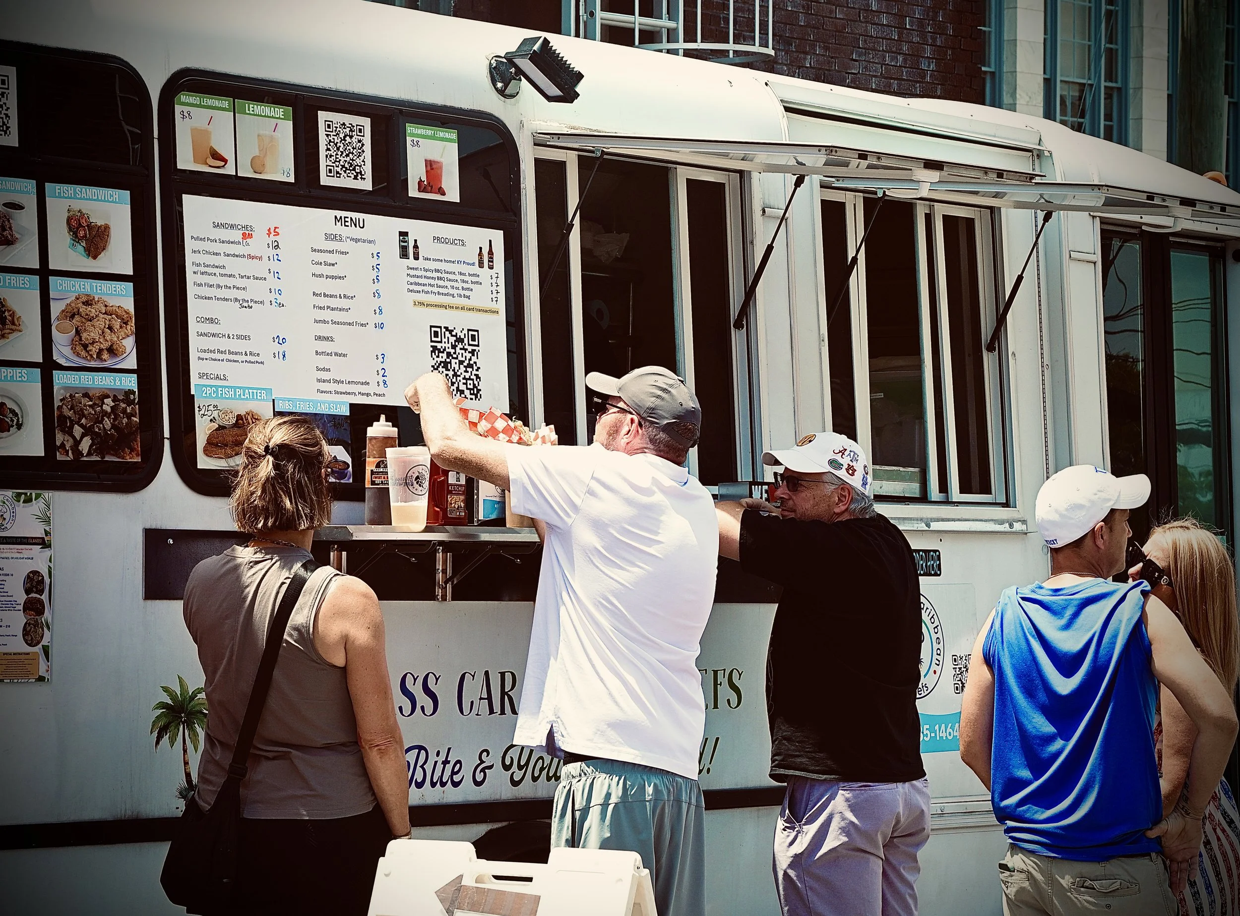 People standing in line at a food truck with a menu displayed on the side, selling sandwiches and fries.