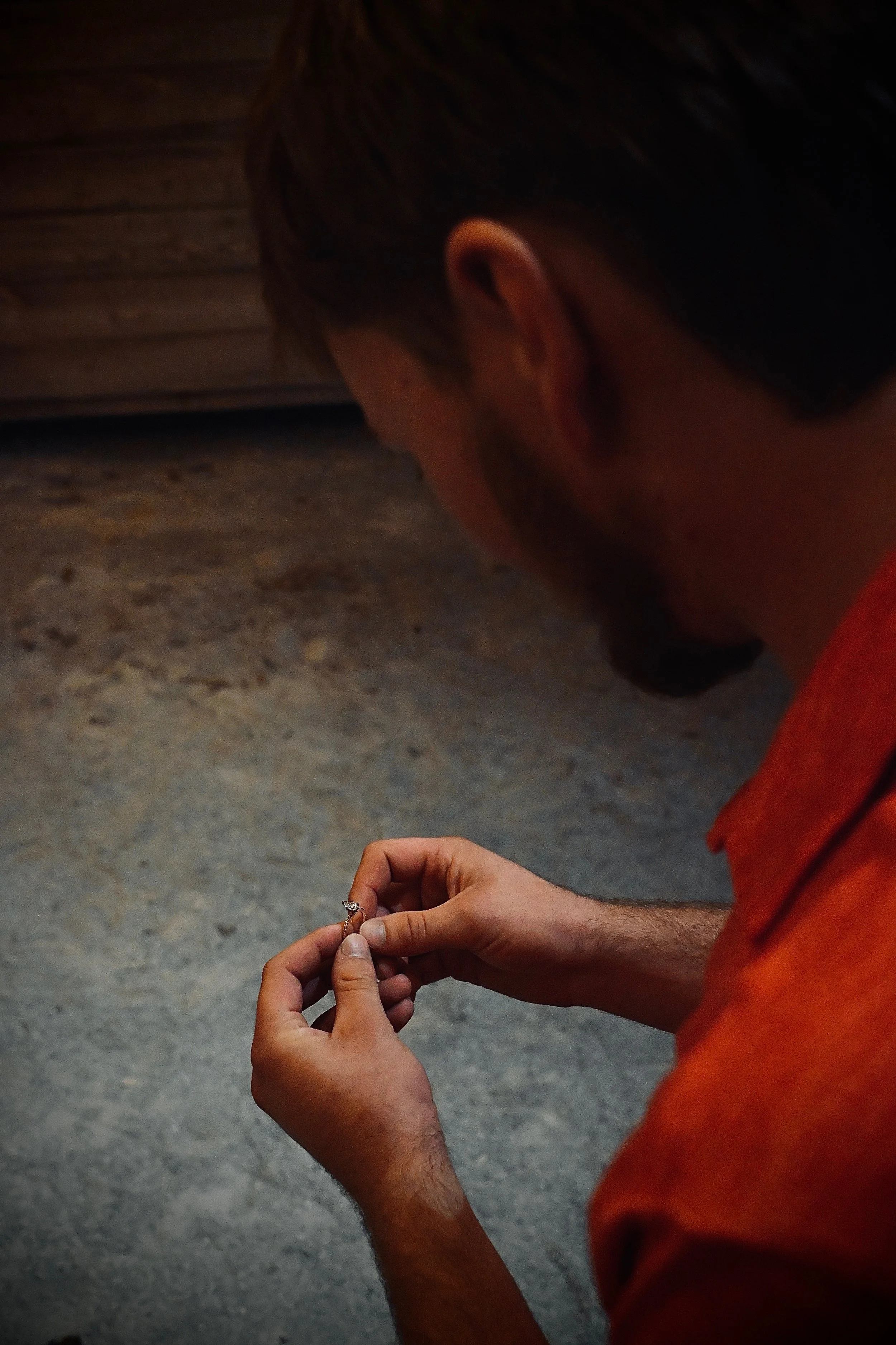 A man, wearing an orange shirt, is holding an engagement ring between his fingers.