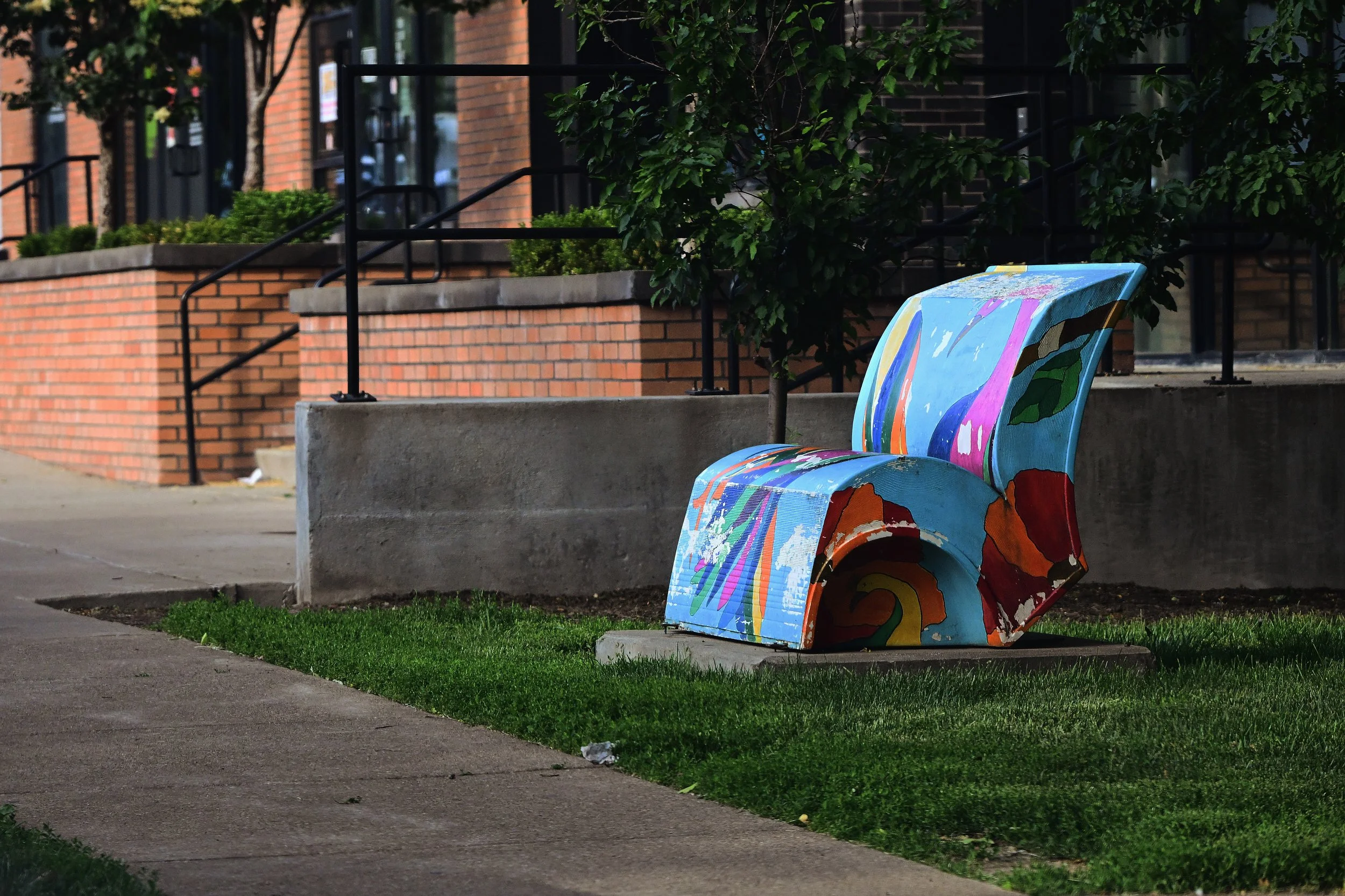 A colorful, painted park bench located on a sidewalk in front of brick apartment buildings with black railings and green bushes in Lexington, Kentucky