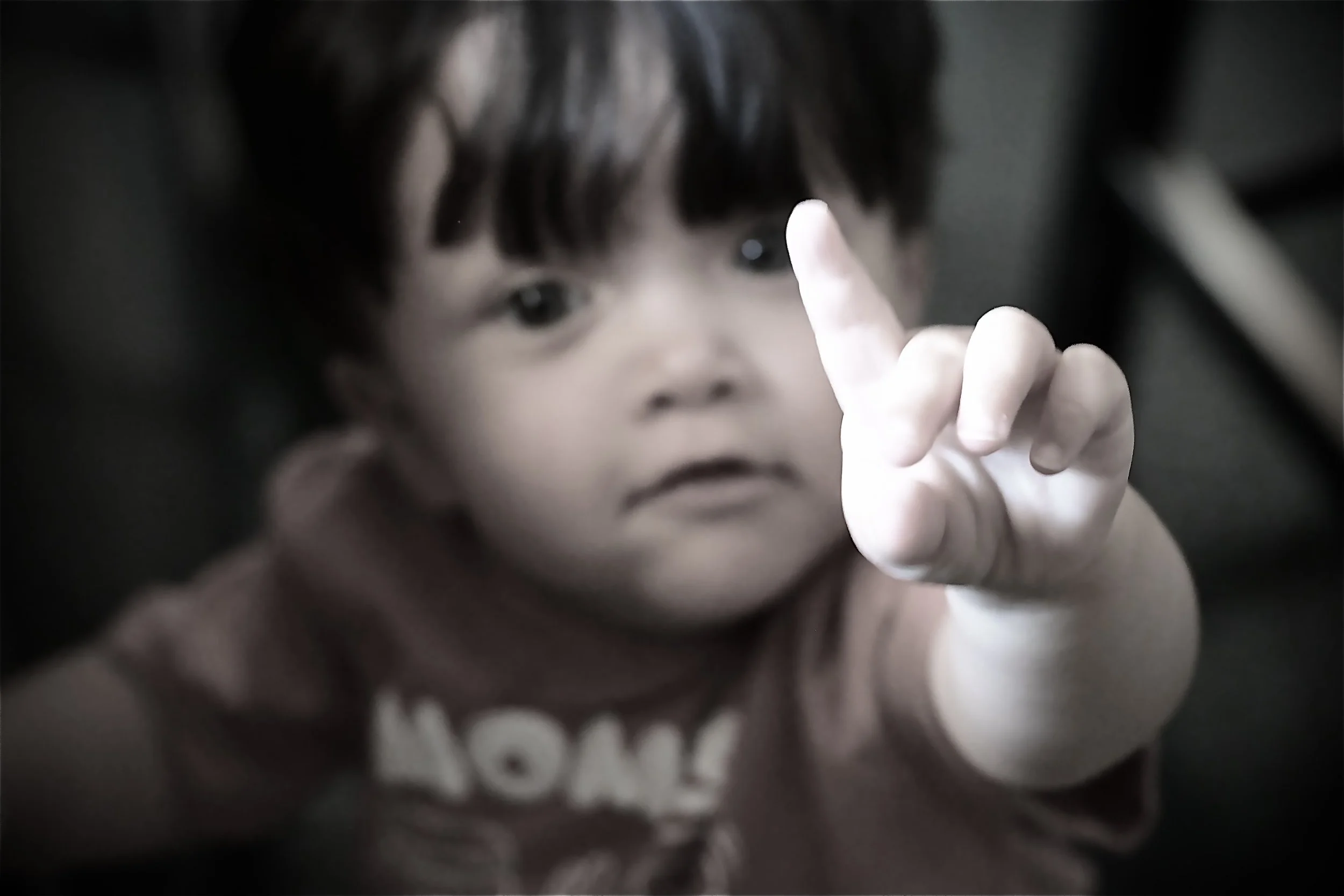 Close-up of a young child's face with dark hair, holding up a finger, with a blurred background.