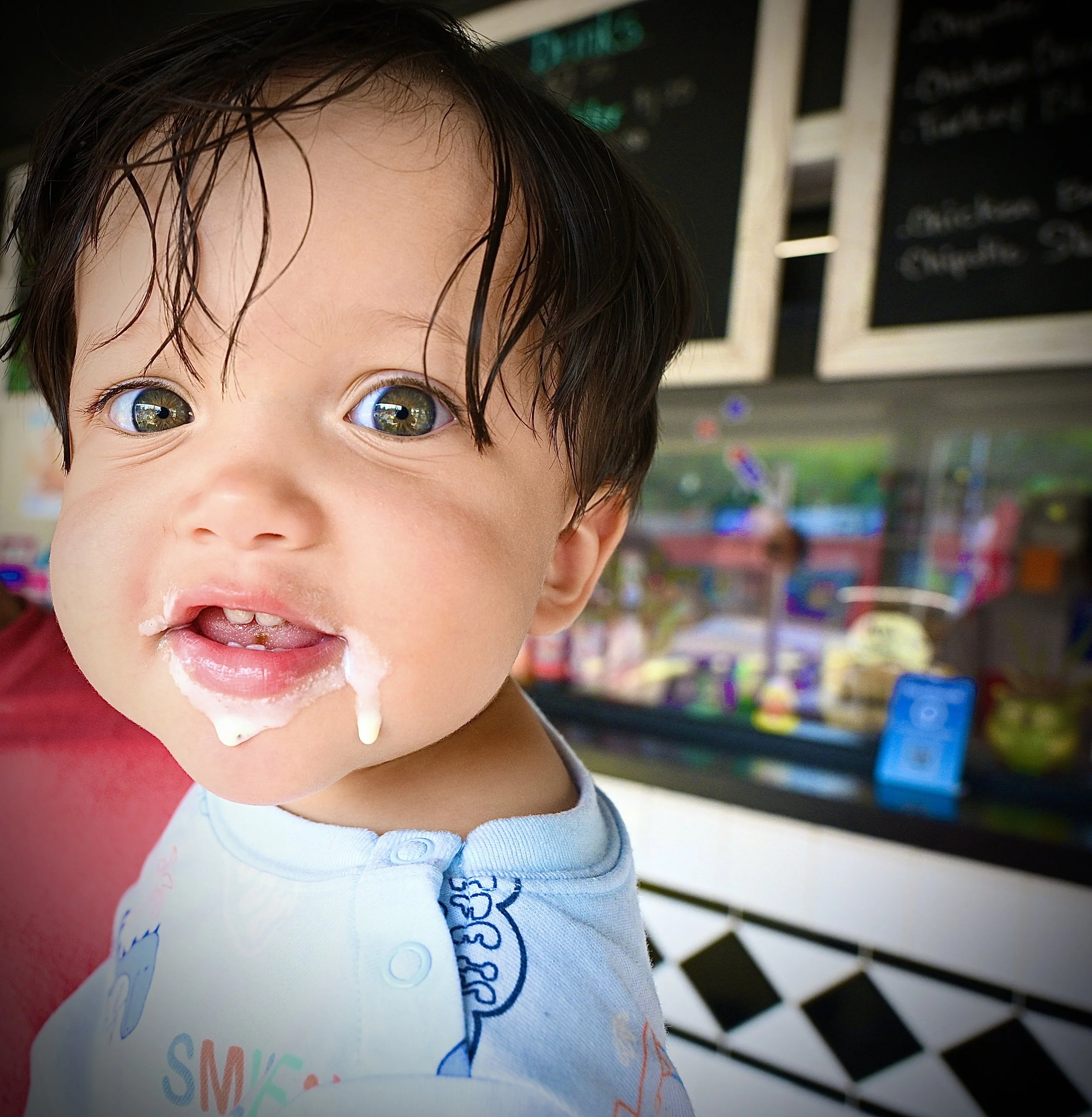 A young child with wet hair, smiling with some food on their lips, wearing a white shirt with a design on it, standing in front of a colorful background with what appears to be a store or market setting.