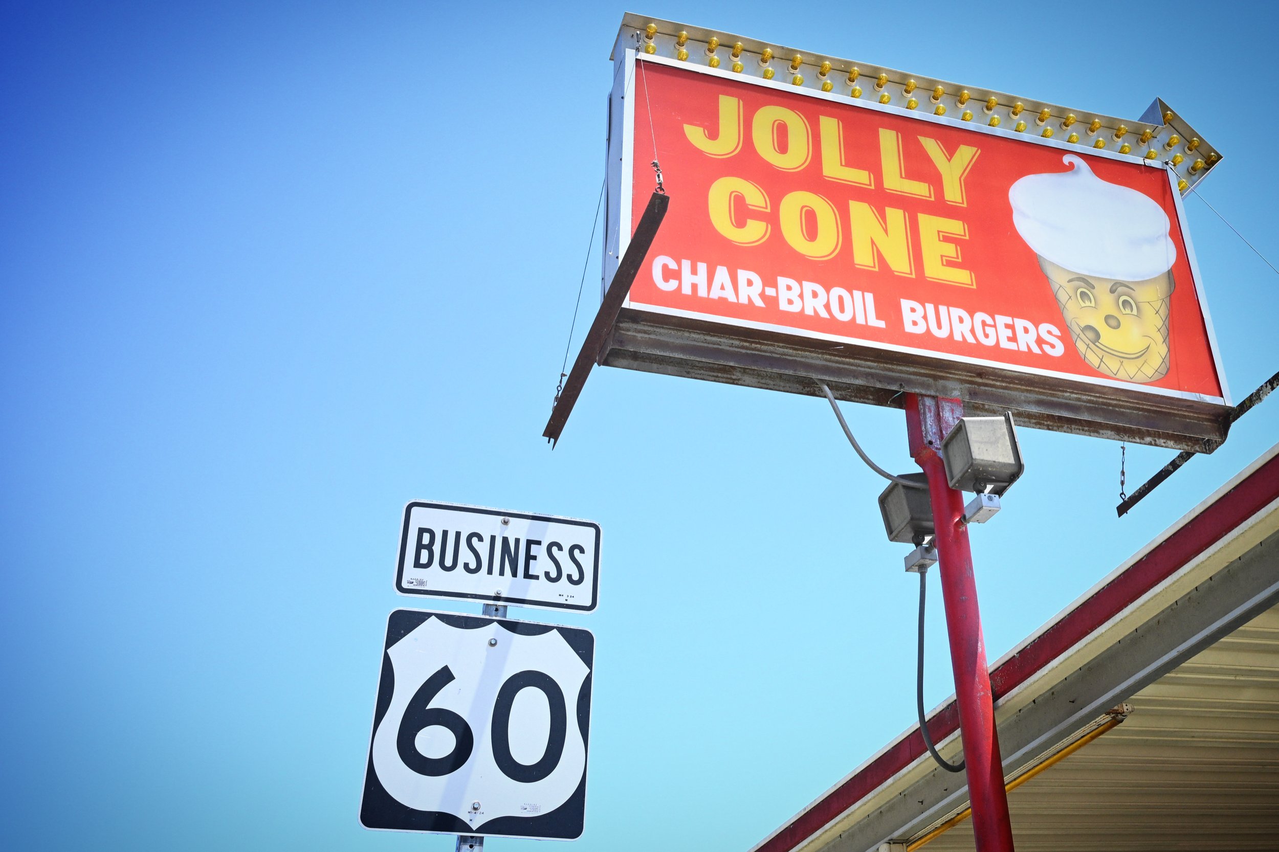 Billboard advertising Jolly Cone, a food stand offering charbroil burgers, with a graphic of an ice cream cone wearing a white hat, against a clear blue sky.