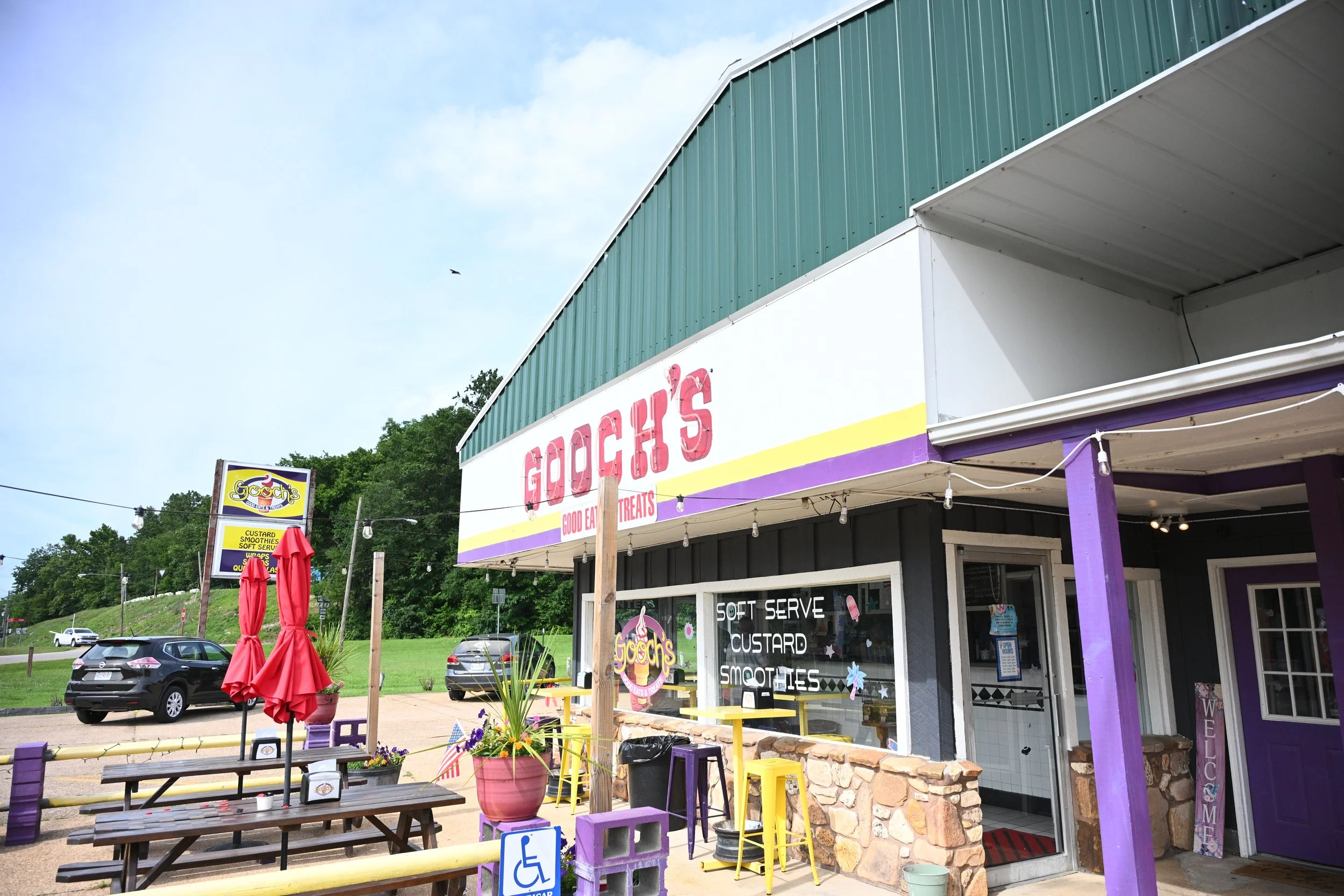 Exterior view of Gooch's dessert shop with colorful outdoor seating, red umbrellas, and a sign advertising custard, smoothies, and soft serve, located on a busy street with cars parked outside.
