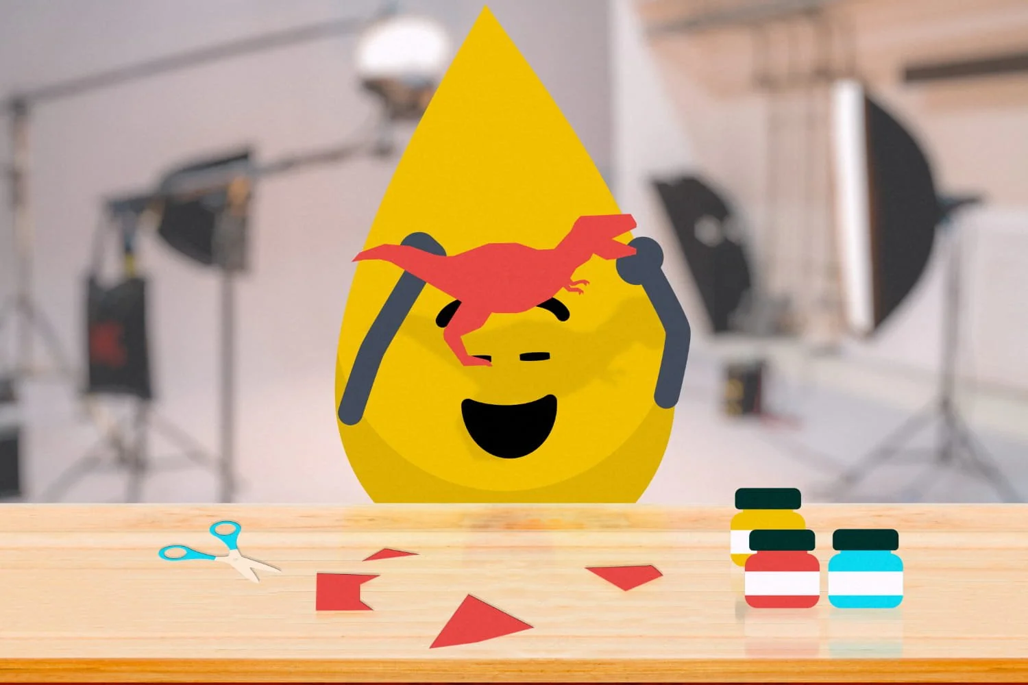 A yellow cartoon water droplet with a smiling face, wearing black sunglasses, with a red paper dinosaur on its forehead. The background shows studio lighting and equipment. On the wooden table, there are small jars of paint, pieces of red paper, and a pair of small scissors.