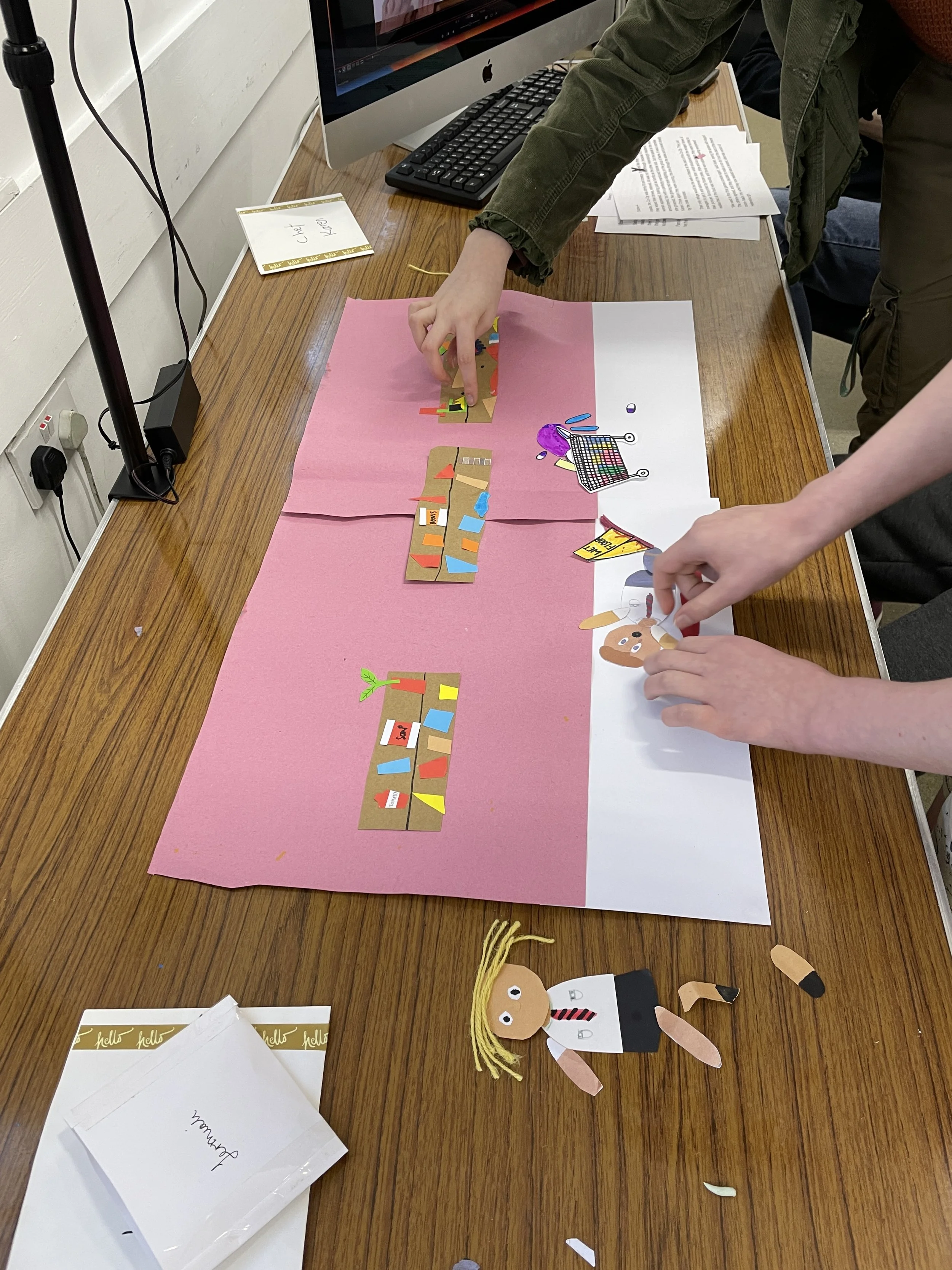 People assembling a collage with colored paper cutouts on a pink and white background, featuring figures and objects. A computer and handwritten notes are visible on the desk.