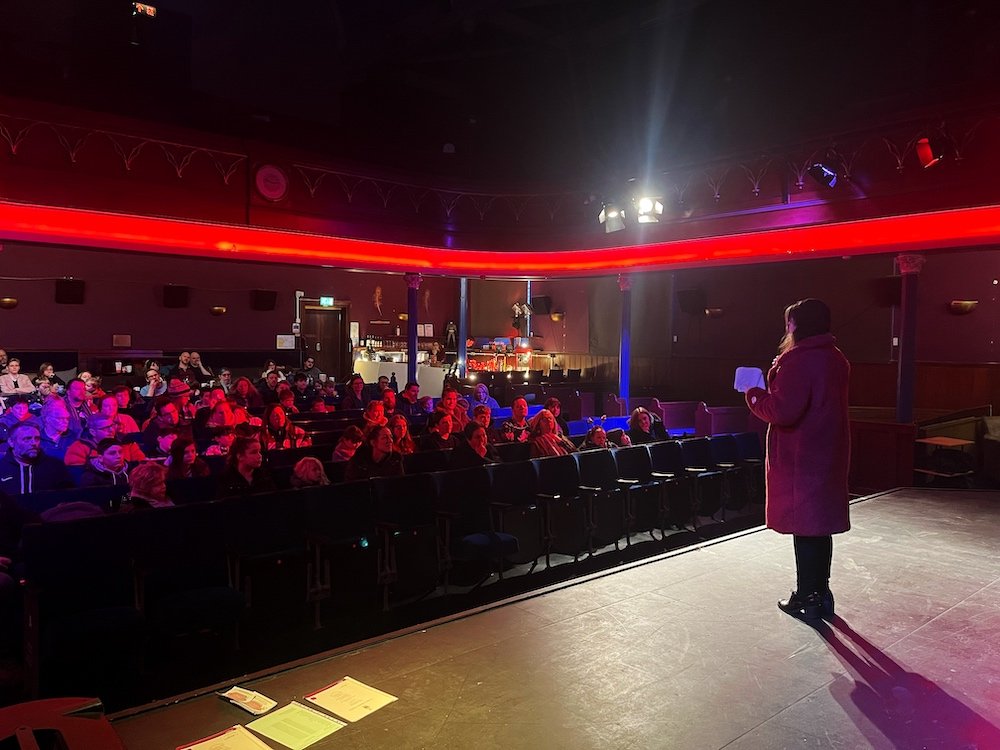Woman gives a speech to a audience in a theatre that is lit red and blue