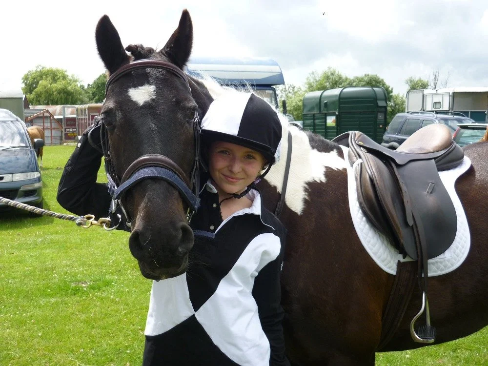 A young girl with a helmet happy beside a black and white horse, in a paddock with vehicles and green trees in the background.