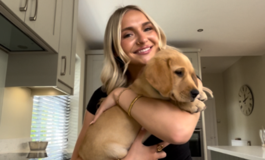 A woman with blonde hair smiling and holding a puppy in her arms inside a kitchen.