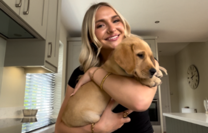 A woman with blonde hair holding a yellow Labrador puppy indoors in a kitchen with white cabinets and a clock on the wall.