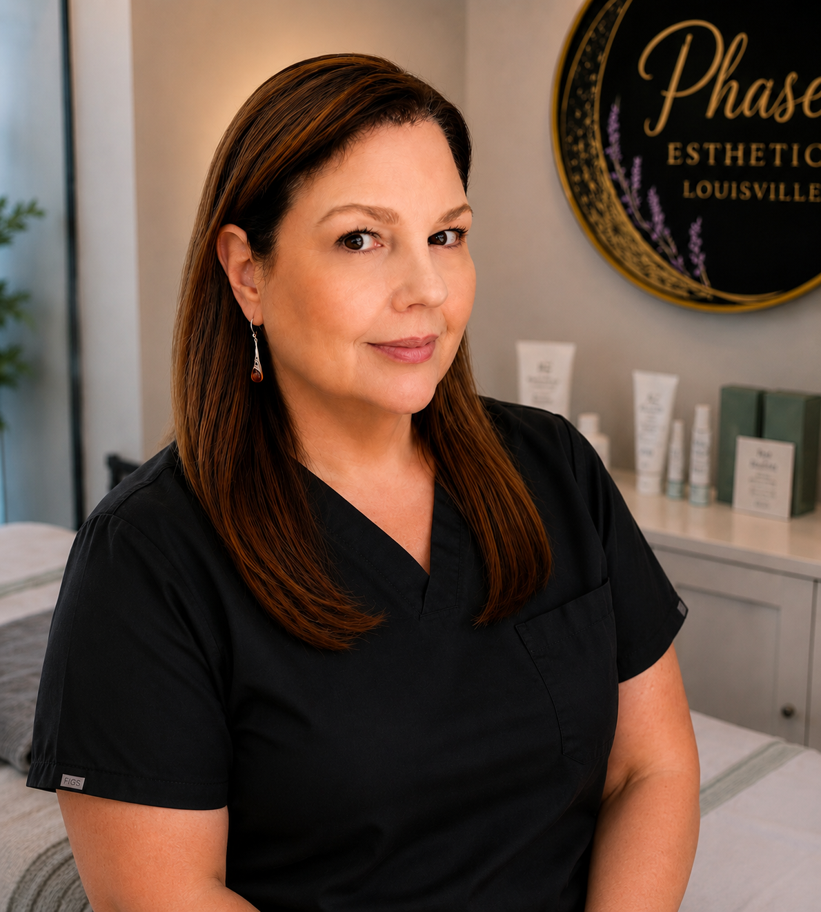 A woman with straight, shoulder-length brown hair wearing a black uniform in a spa or skincare clinic. She is smiling softly and looking slightly to the camera's right. Behind her, there are skincare products on a counter and a round sign that reads 'Phase Esthetics Louisville'.