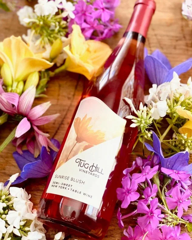 A bottle of Tuc Hill Vineyards Sunrise Blush semi-sweet table wine surrounded by colorful flowers on a wooden surface.