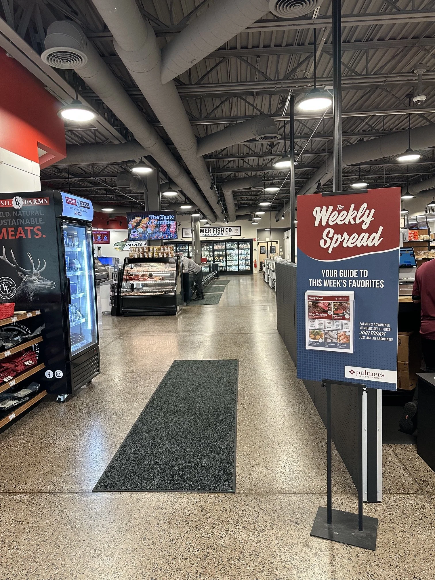 View of the interior of a grocery store with red and blue signage, display cases, and a person shopping in the background. A sign on the right reads 'Weekly Spread' with a guide to weekly favorites.