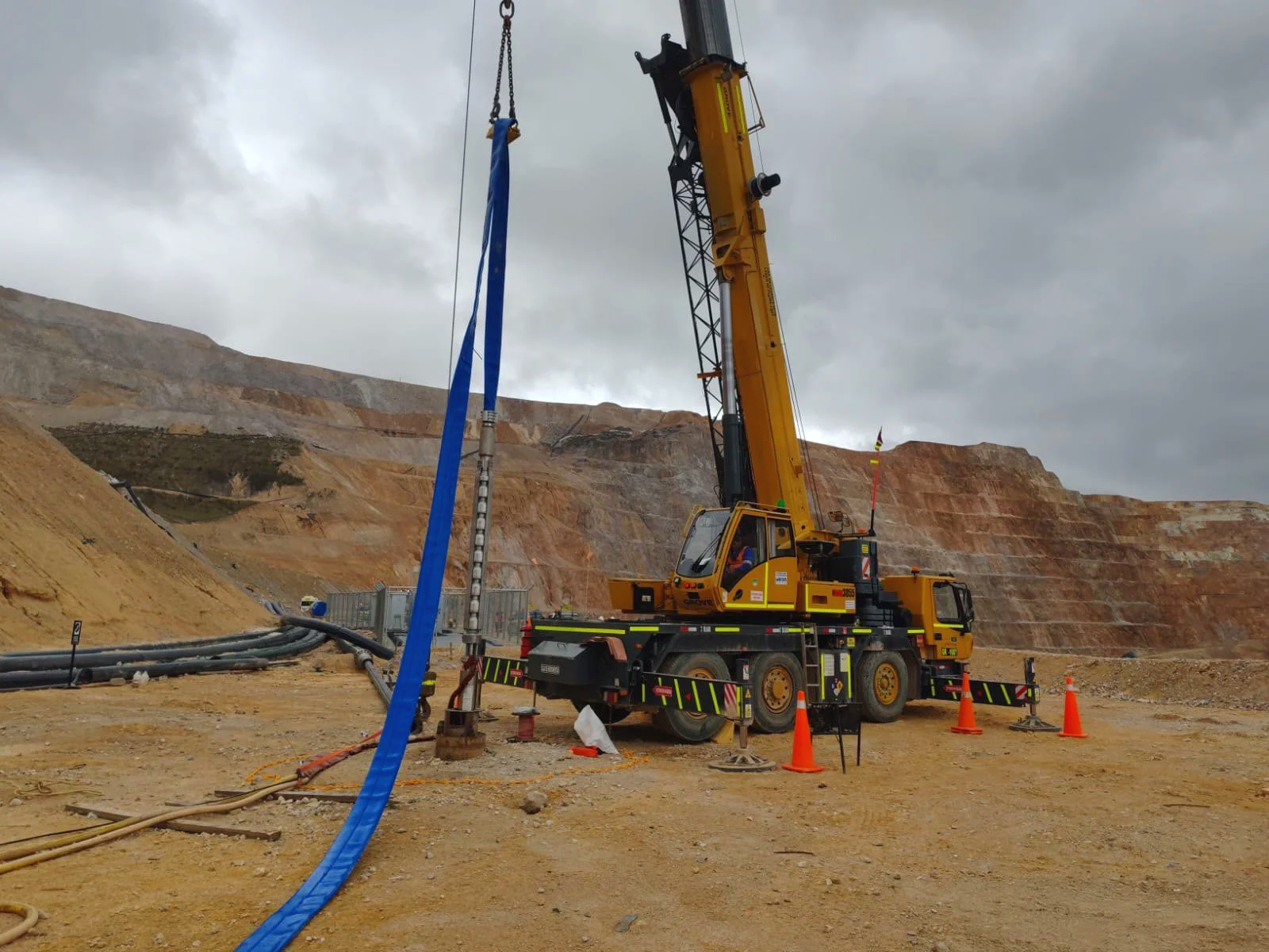 A large yellow crane is at a construction site operating toward the ground with cables, hoses, and orange safety cones surrounding it. The background features a rocky hillside under cloudy skies.