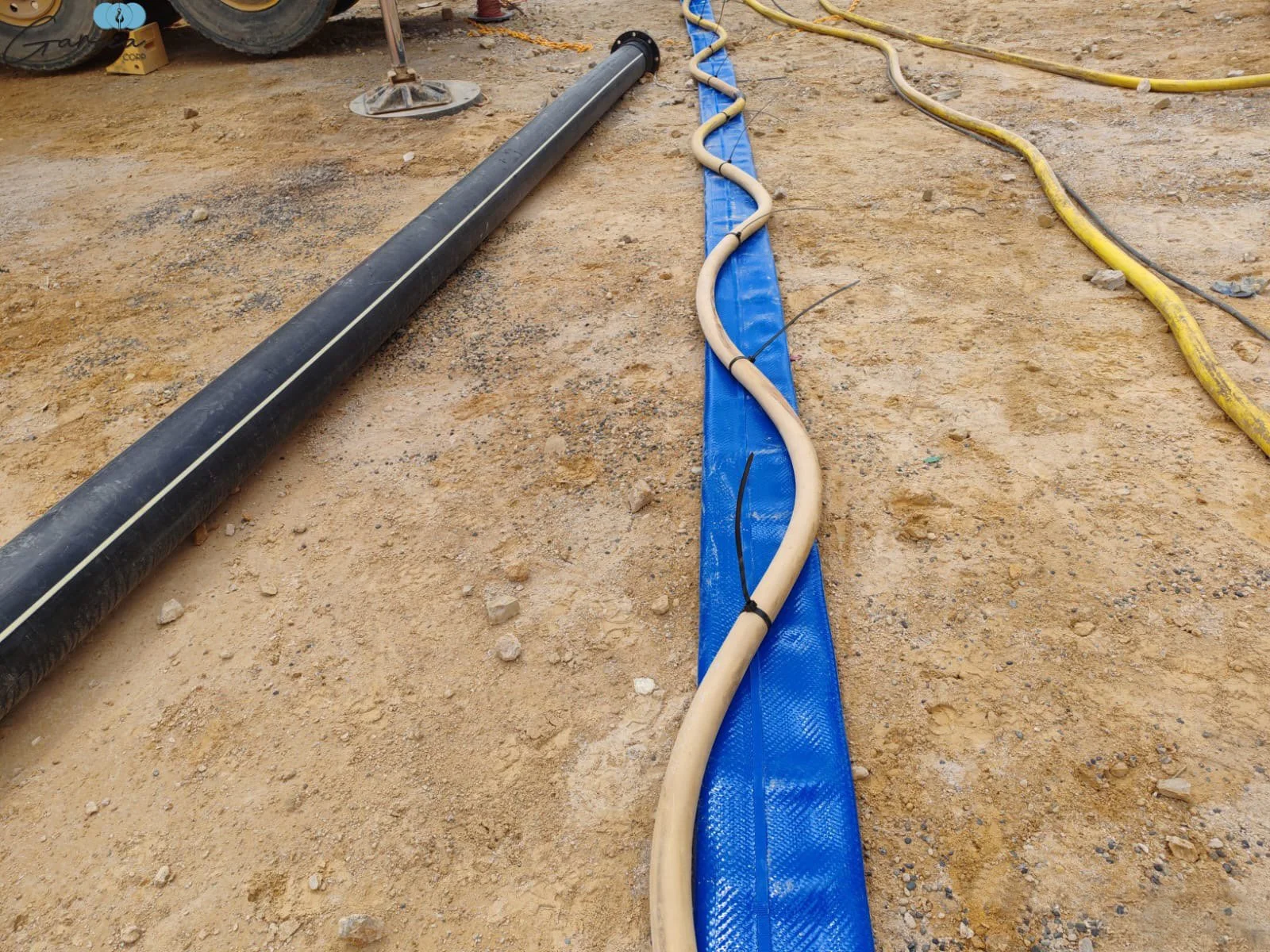 Construction site with black pipe, yellow and beige conduit on blue ground sheet, and yellow hoses on dirt ground.