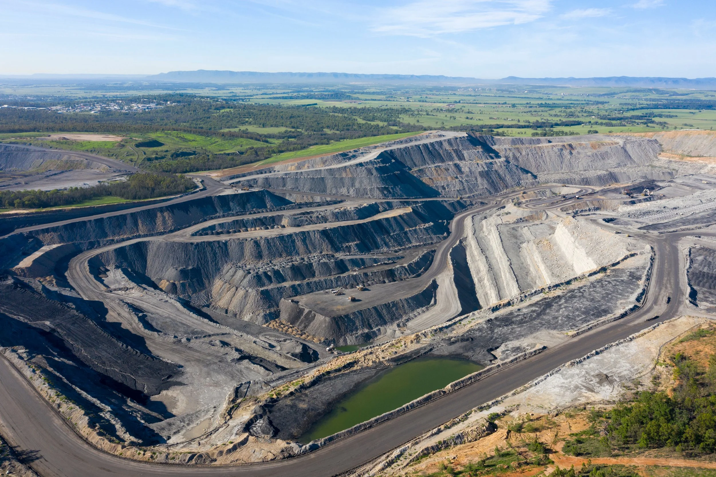 A large open-pit quarry with terraced steps and a green water pond at the bottom, surrounded by forested areas and distant mountains under a blue sky.