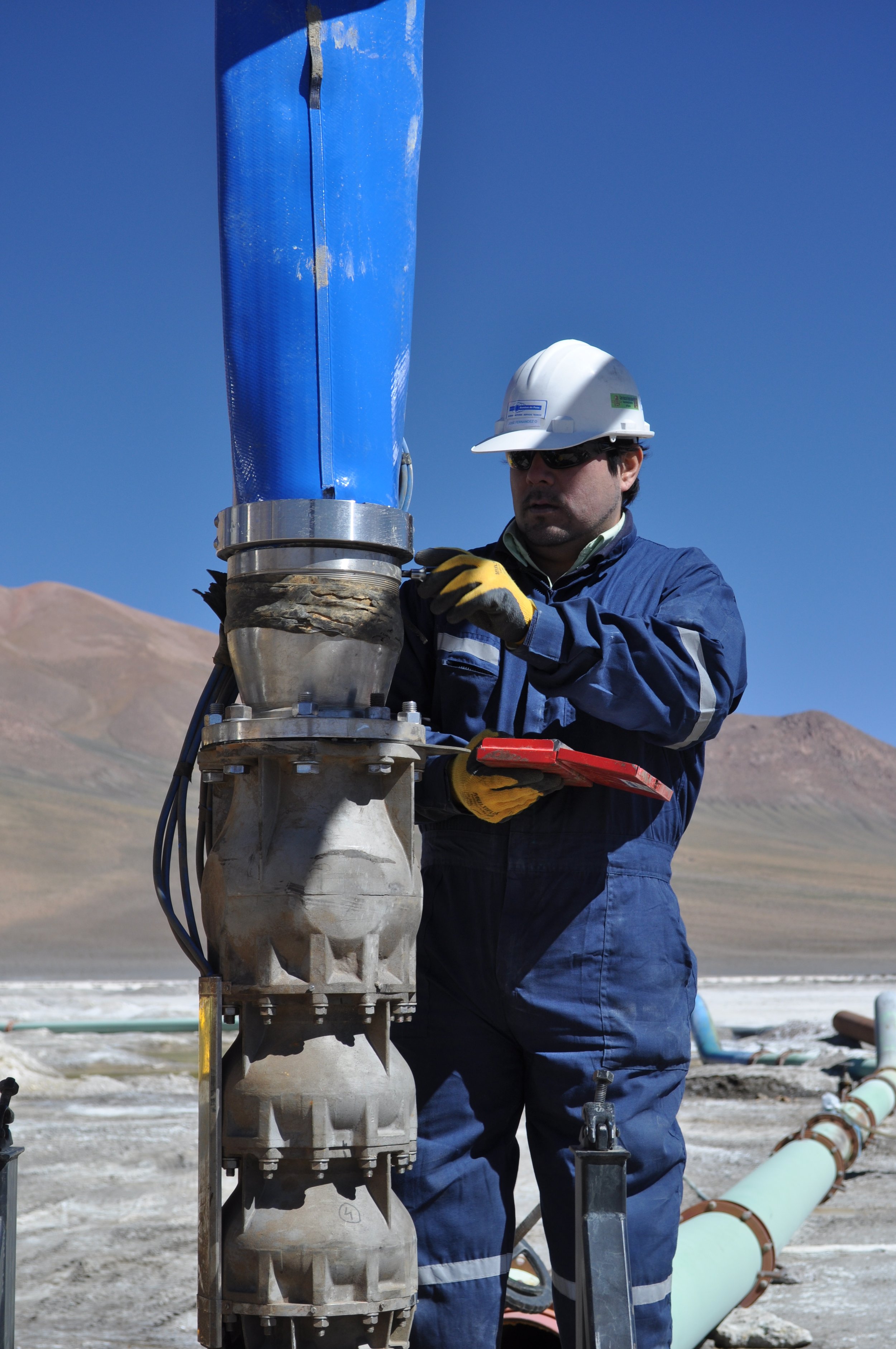 A worker wearing a white safety helmet, sunglasses, and blue coveralls is inspecting industrial piping outdoors, with mountains and clear blue sky in the background.