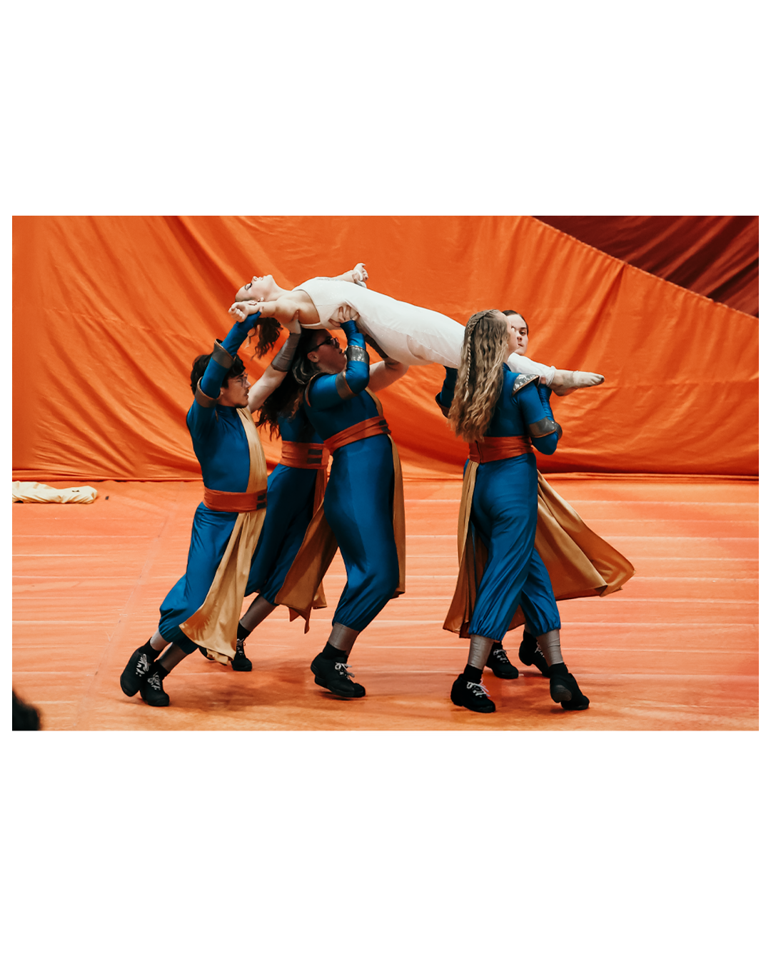 A group of performers in blue and gold costumes lift a person in a white costume during a dance or theatrical performance on a stage with an orange background.