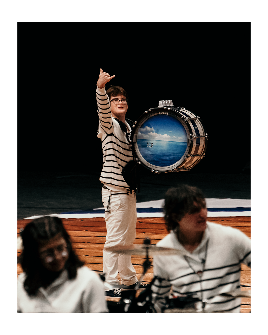 A young man holding a large drum with a painted ocean scene, standing on a wooden stage, with other people in the foreground.