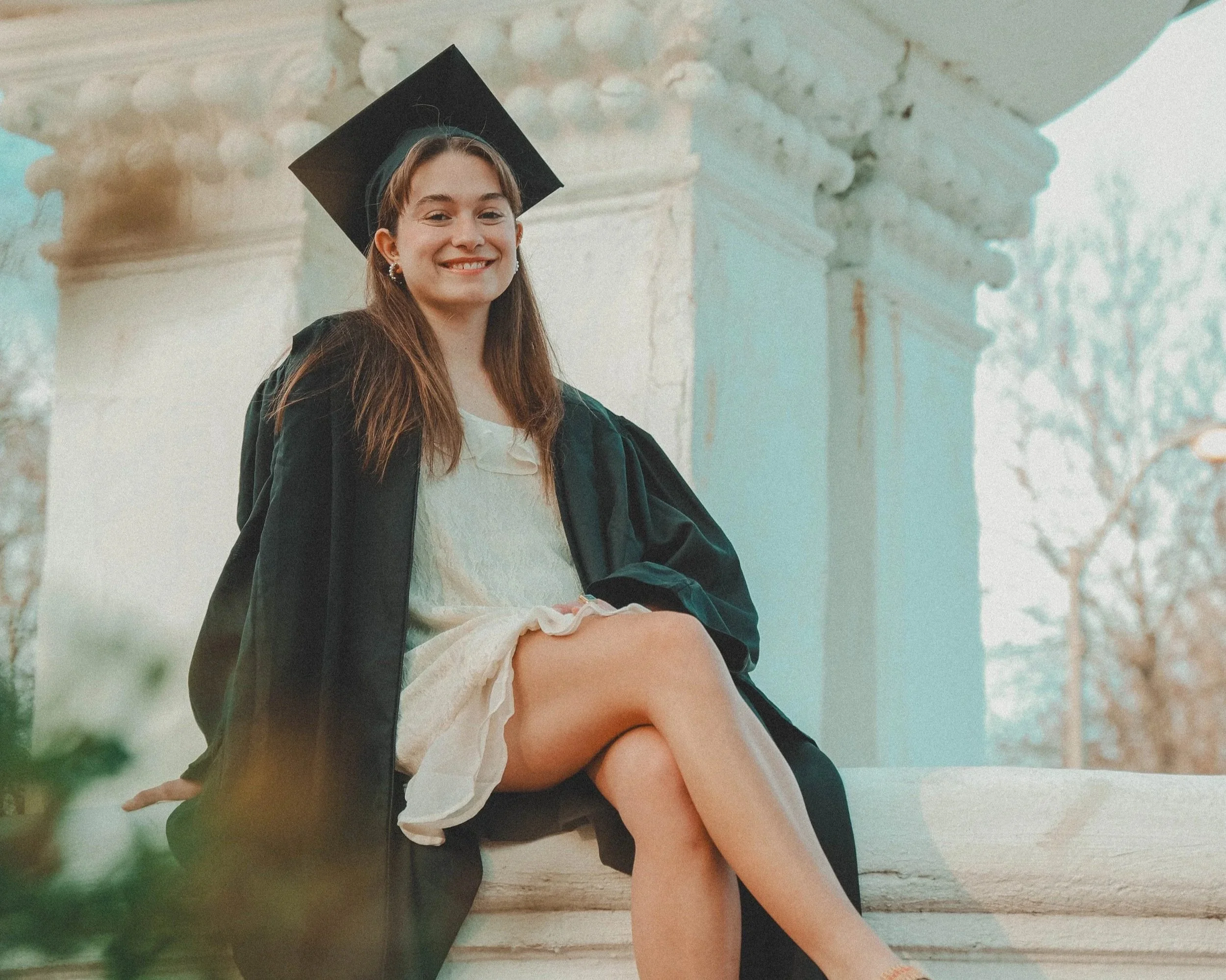 A young woman in a graduation cap and gown sitting on steps outside, smiling, with one leg crossed over the other, wearing sandals, during daytime with a white structure in the background.