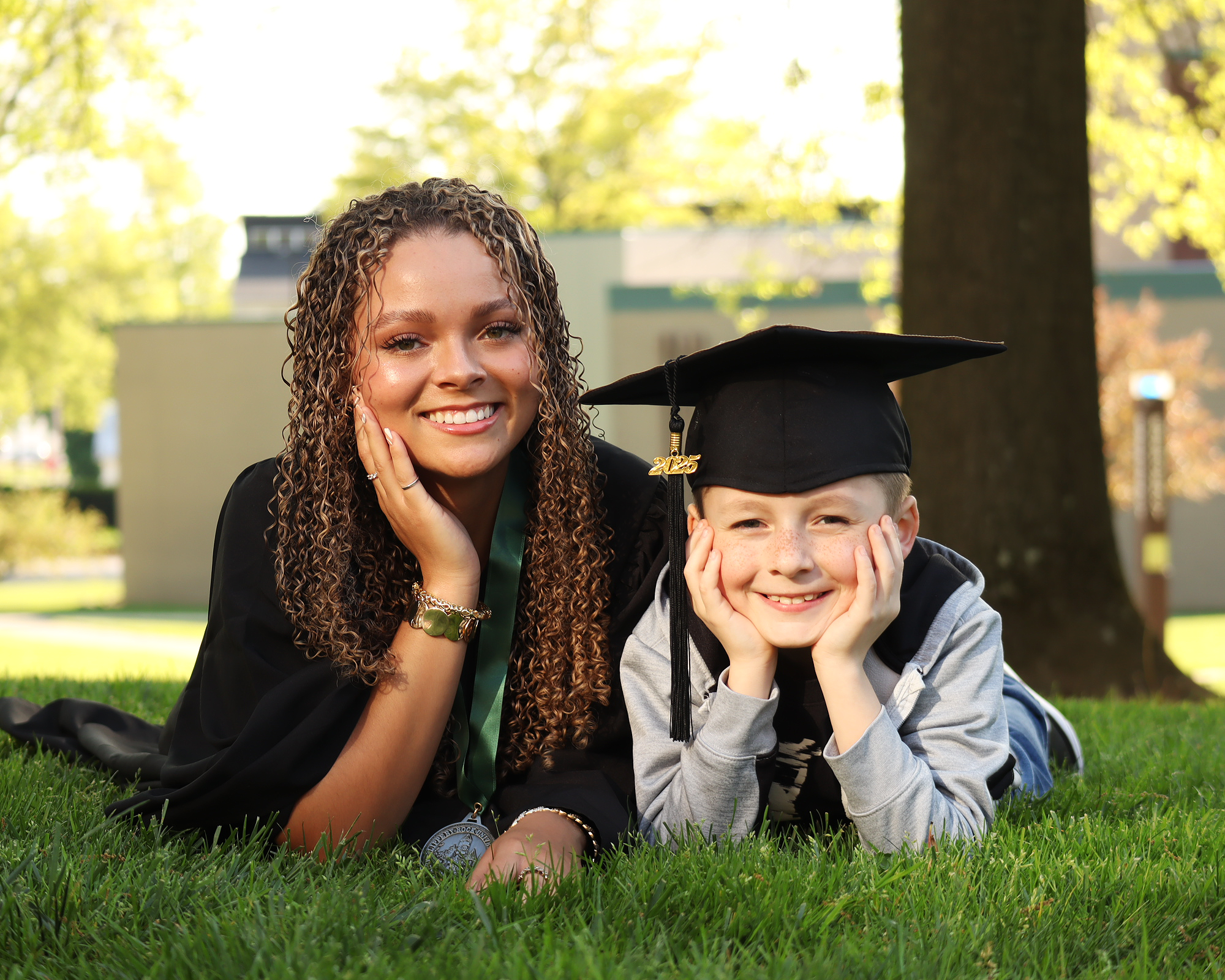 A woman with curly hair and a young boy in a graduation cap and gown, lying on the grass outdoors with trees and buildings in the background, celebrating graduation.